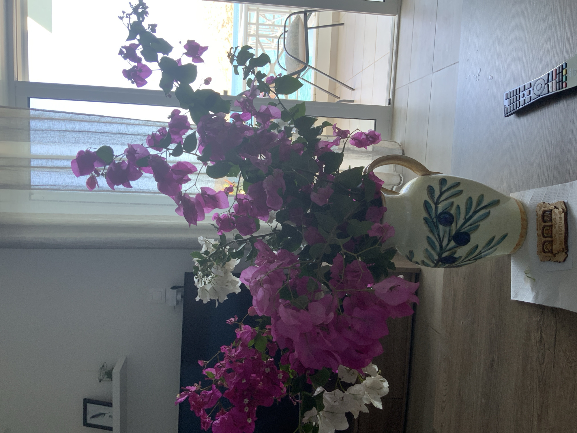 A still life image captured indoors shows a bouquet of pink and white bougainvillea flowers in a decorative ceramic vase. The vase features a green olive branch motif. The vase sits on a wooden surface, likely a table, in front of a large glass door that opens to a balcony or patio. Beyond the glass, a view of the sea and a chair with a cushion can be seen. The room appears to be a living space, with a dark furniture piece and a shelf with framed artwork visible in the background. A remote control rests on the wooden surface near the vase. The overall impression is a peaceful domestic setting with a view of the coast. The image is likely taken in Agios Nikolaos Municipal Unit, Greece, given the presence of bougainvillea and the coastal scenery.