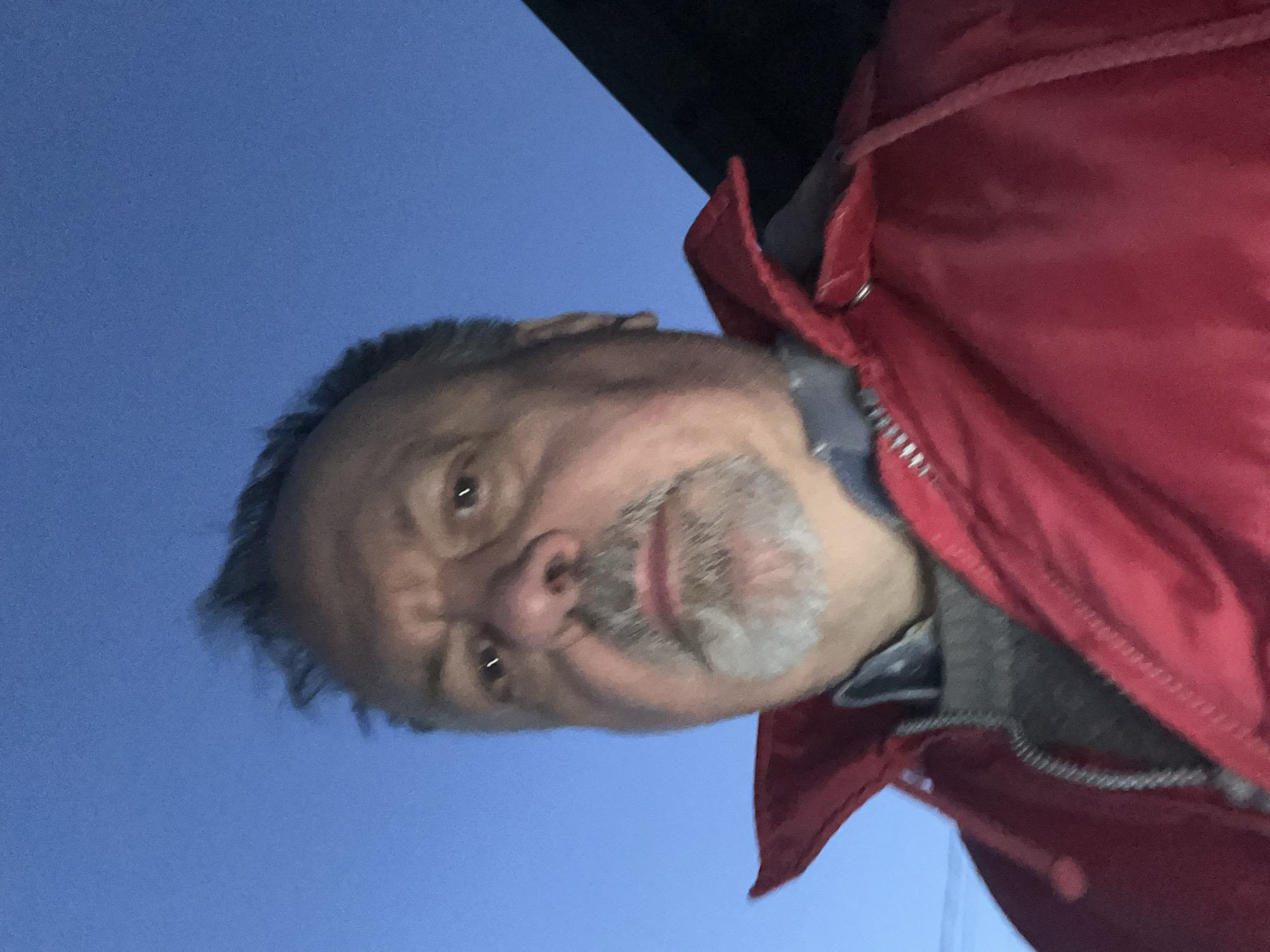 A middle-aged man with graying hair and a gray and white beard is wearing a red jacket. The background shows a blue sky. The photo was taken from a low angle, looking up at the man. The location is unknown; there is no indication it is Podolsk, Russia.
