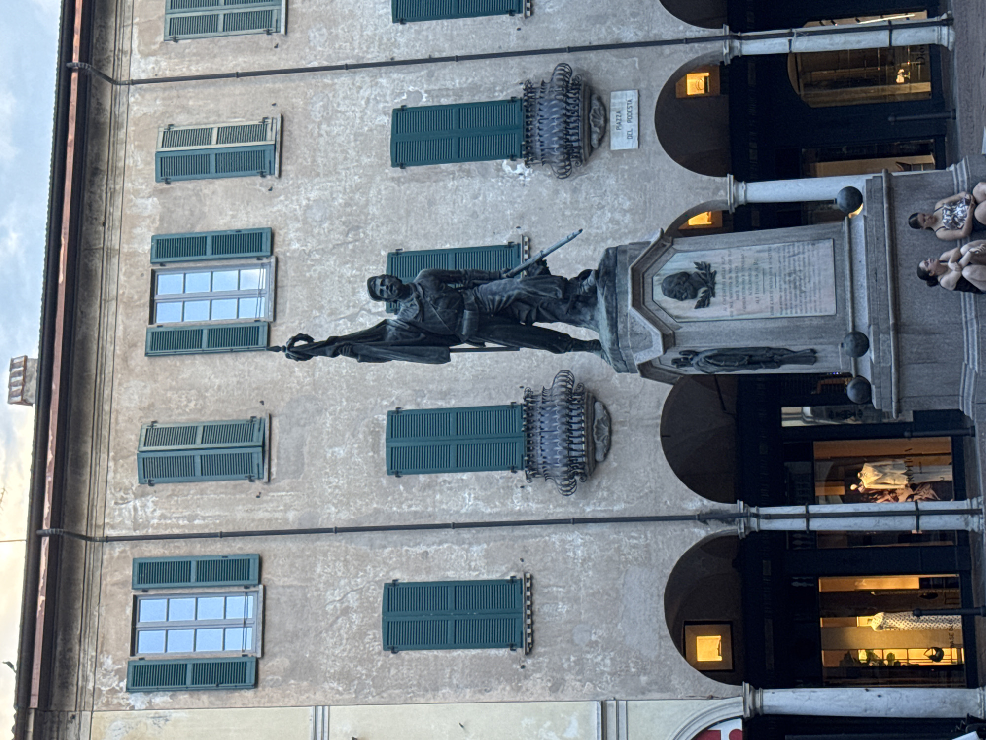 The image shows a statue in a public square in Varese, Italy, identified by a plaque on the building as "PIAZZA DEL PODESTA". The statue depicts a standing male figure holding a flag and a rifle, dressed in what appears to be military attire. The statue is on a large, ornate pedestal with inscriptions and bas-reliefs. In front of the pedestal, two young women are sitting, one looking at her phone and the other looking forward. The background features a building with multiple windows, many with green shutters, and arched storefronts. The architecture suggests a European city.