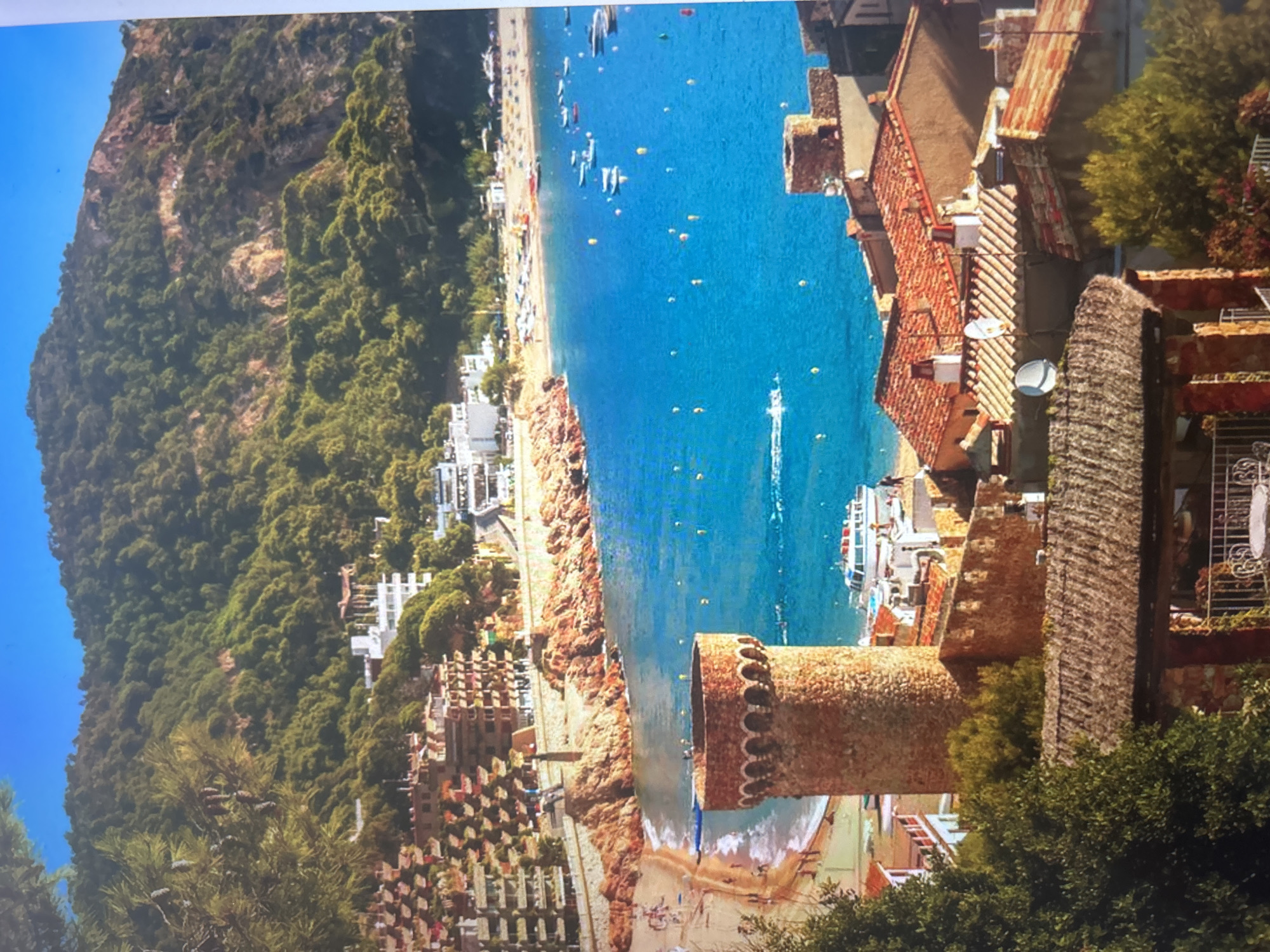 This media file presents a vibrant, high-angle view of a picturesque coastal town, likely situated along a Mediterranean coastline. The scene is dominated by natural beauty and traditional architecture, bustling with leisure activities.
On the left, a steep, verdant mountain covered in dense green trees and foliage rises majestically, with some exposed reddish-brown rock faces visible. The sky above is a clear, bright blue, indicating sunny weather.
The central part of the image features a stunning bay with crystal-clear turquoise waters. Numerous boats, including sailboats, yachts, and smaller vessels, are scattered across the water, some moored and others potentially sailing. Colorful buoys mark various spots in the bay. A white motorboat can be seen in the distance, leaving a visible wake. A sandy beach curves along the shore, lined with sunbathers, umbrellas, and what appear to be beachfront buildings or facilities.
To the right, the town's architecture features prominently. Buildings with traditional terracotta tiled roofs and stone or light-colored facades are built into the hillside, cascading down towards the sea. A striking circular stone tower or fortification, with arch-shaped openings near its top, stands prominently on the coast, near the beach. Lush green trees and vegetation are abundant, interspersed among the buildings and along the coastline.
The overall impression is that of a popular and scenic holiday destination, teeming with activity during what appears to be a bright, clear day. The time of day is clearly daytime, likely mid-morning to afternoon, given the bright illumination and lack of long shadows. There is no visible text in the image.