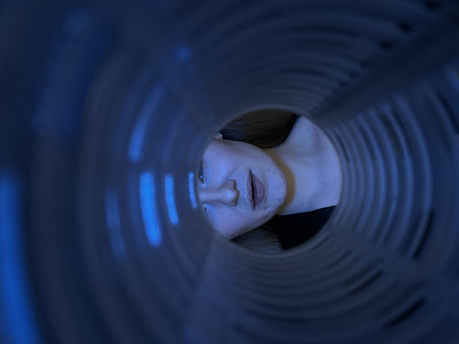 A woman's face appears at the end of a cylindrical structure. Her head is tilted down and to the left, with her face centrally framed by the opening. Blue light illuminates her face and the interior of the structure, casting a cool tone. Ribbed patterns line the interior, leading the eye toward the face. The outer edges of the structure are blurred, creating depth.