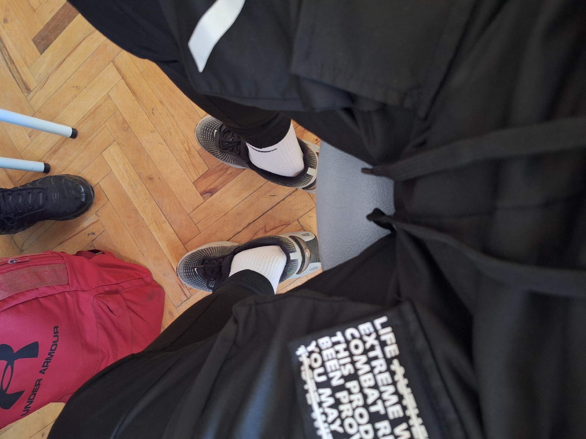 An elevated point-of-view shot shows a person's legs and feet, clad in black athletic pants and white socks, with black and gray athletic shoes on. The person is seated on a gray upholstered surface, with their legs spread apart. To the left of the seated person, a red Under Armour backpack is visible, resting on a polished wooden floor. Further to the right, partially obscured by the seated person's leg, is a black athletic shoe positioned on the same wooden floor. Two white legs of a table or chair are also visible on the wooden floor in the upper right portion of the image. The setting appears to be indoors.