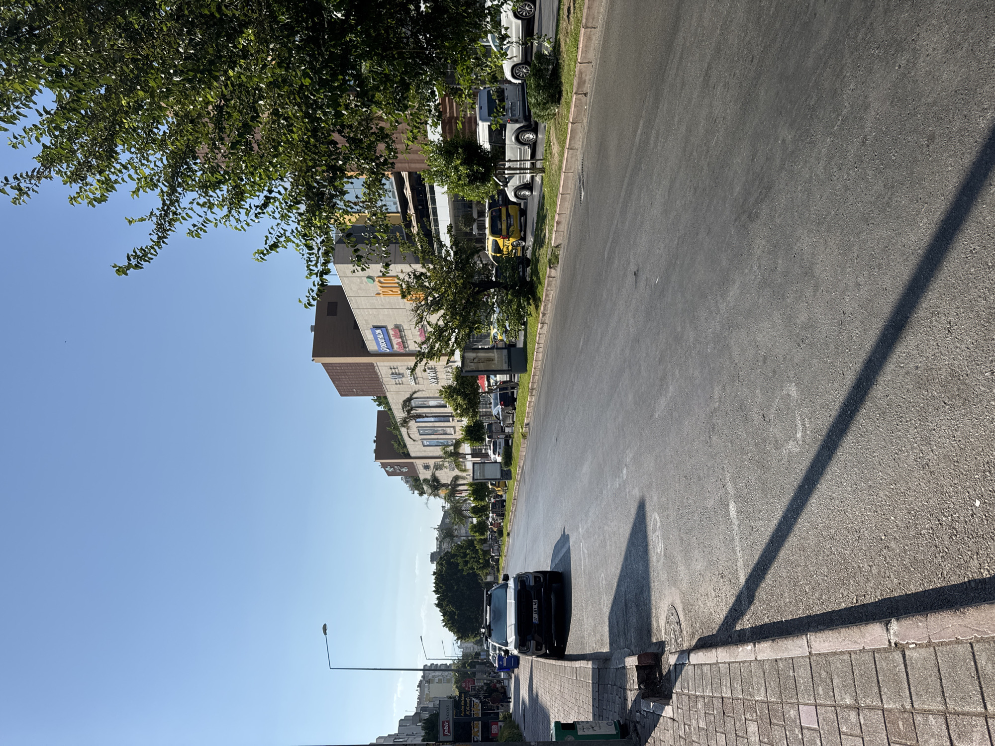 This is a medium-angle, eye-level, full-shot photograph of a street scene in Antalya, Turkey, on a clear, sunny day. The image is dominated by a wide, empty asphalt road that curves gently to the right. On the left side of the road, there is a sidewalk paved with square tiles, bordered by a raised curb. A black SUV is parked on the street, facing forward, with its tires just touching the curb. A street lamp stands on the sidewalk, casting a long shadow across the road.
On the right side of the road, there is a row of trees and then a large, multi-story building complex with a modern facade, featuring glass windows and balconies. Several vehicles, including a yellow taxi and a white van, are visible parked along the side of the road in front of the building. The architecture suggests a commercial area, possibly a shopping center or office complex. The background shows more buildings and trees, with a clear blue sky overhead. The overall impression is that of a bustling urban environment on a bright day.