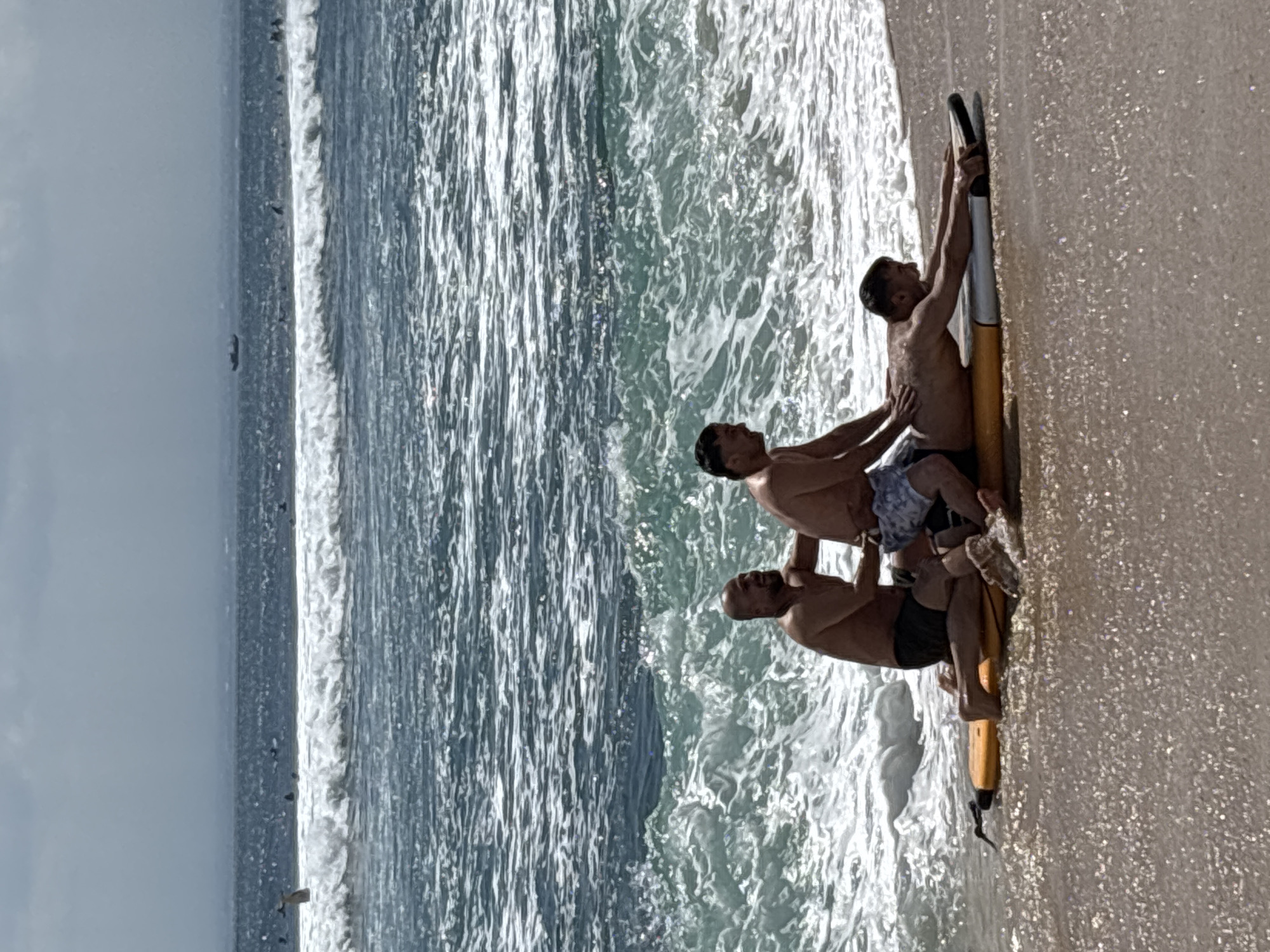 The image captures three adult males at the water's edge on a beach in Kuta Selatan, Indonesia, during daylight hours. A male figure lies prone on a yellow and white surfboard positioned on the wet sand, facing right with his arms extended forward on the board. Immediately behind him, a second male kneels on the surfboard, his hands placed on the back of the prone male. To the left of the second male, a third bald male kneels on the wet sand, his hands also in contact with the backs of the other two individuals. All three are shirtless and wear swim shorts. The background features the ocean with multiple lines of white-capped waves. In the mid-distance, numerous small dark figures are visible in the water, consistent with bathers or surfers. A dark vessel is present on the horizon. The sky above is partially overcast.