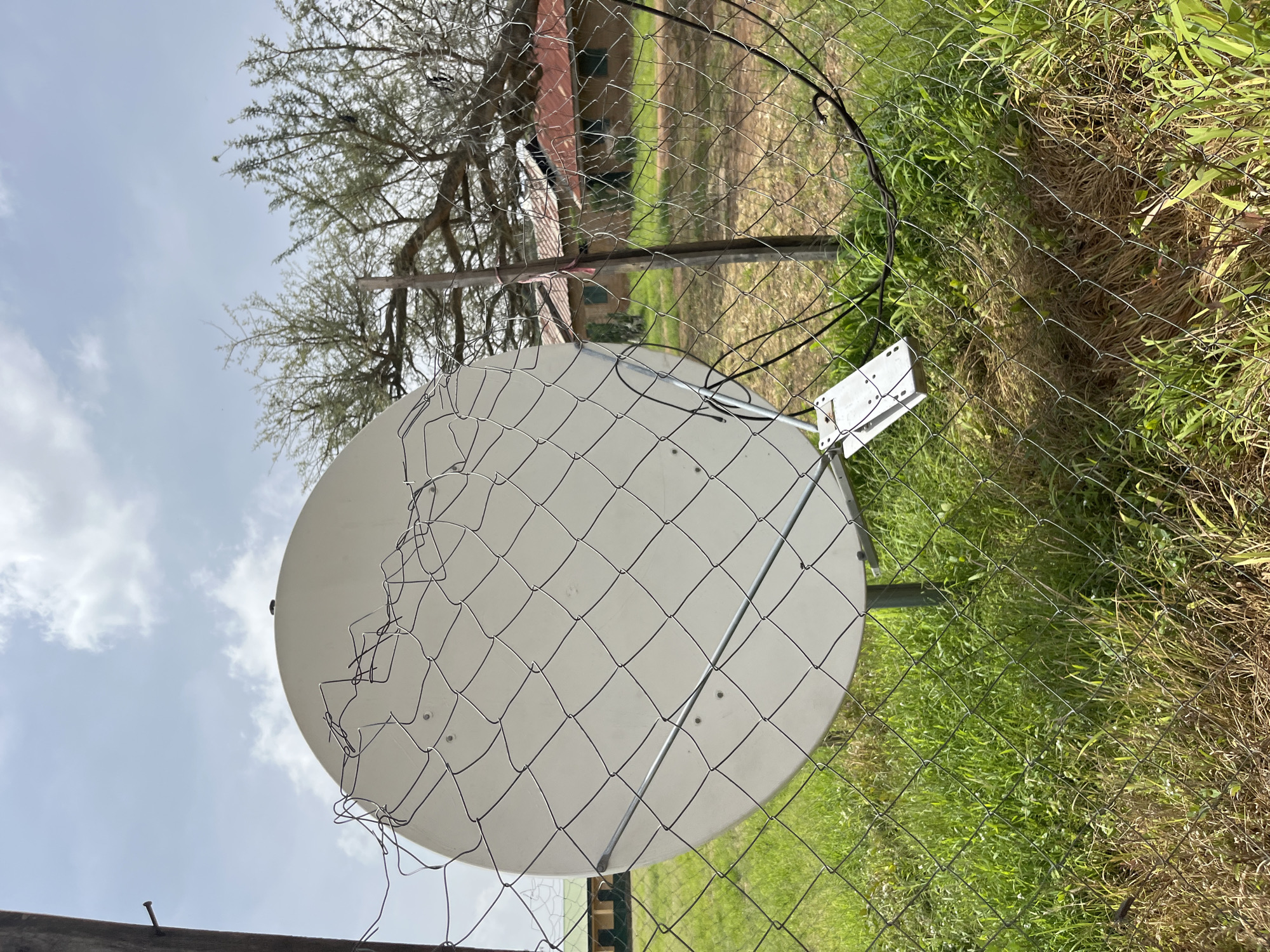 In Bukuru, Nigeria, a white satellite dish is mounted on a metal pole, partially obscured by a chain-link fence. The dish's surface is clean, and it is attached to a bracket. A black cable runs from the dish and is secured to the fence. The fence itself is made of wire mesh and appears somewhat damaged with bent sections. Behind the fence, overgrown green grass and dry brush are visible. In the background, there are a few buildings with red-tiled roofs and green-painted window frames. Trees with sparse foliage are also present. The sky is overcast with scattered clouds.