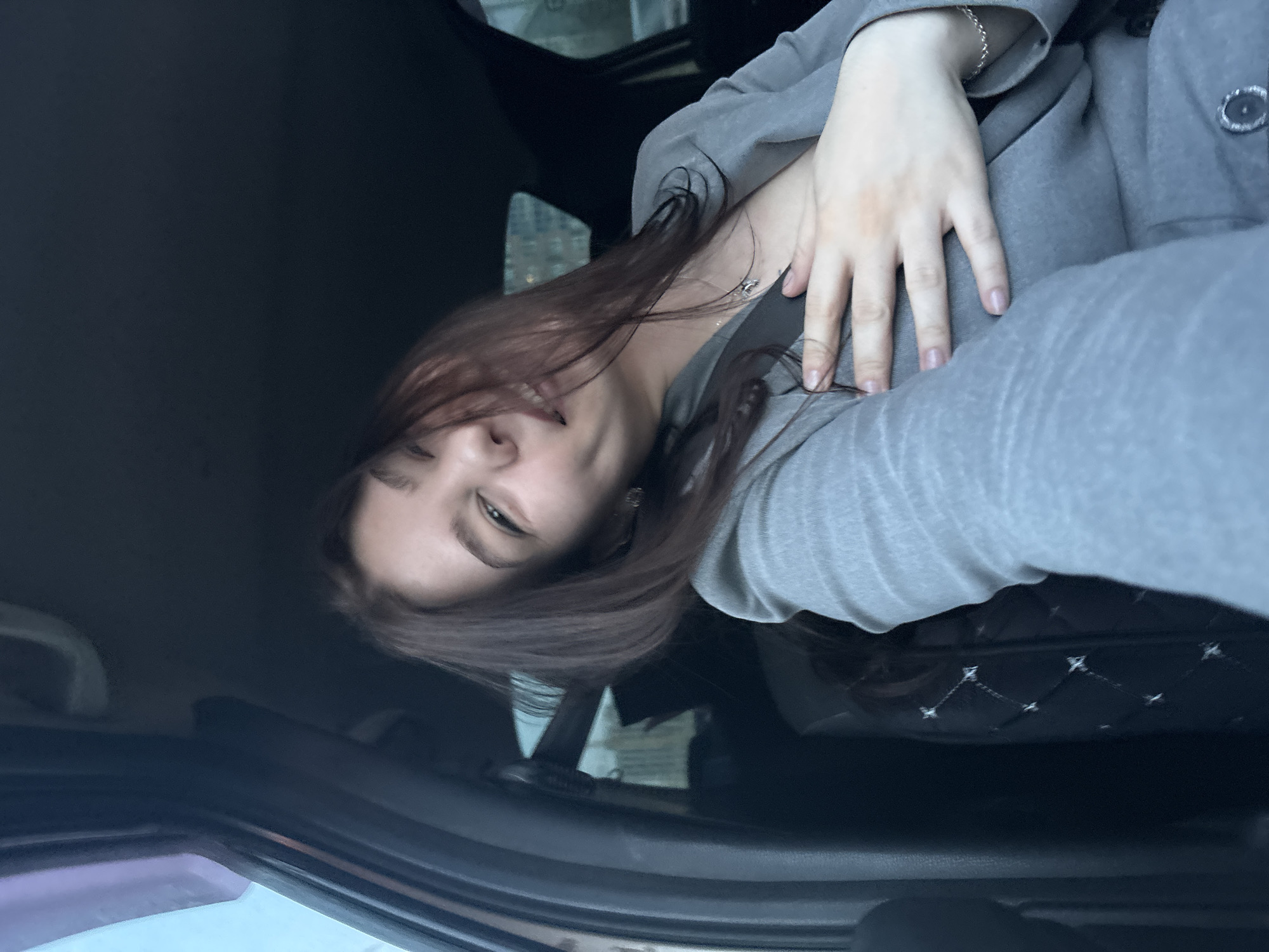 A woman is taking a selfie inside a car in Perm, Russia. She has medium-length brown hair, fair skin, and is wearing a gray blazer and a silver bracelet. She is holding her hand on her chest and smiling slightly. The interior of the car has dark upholstery with a quilted diamond pattern. The car is parked, and a glimpse of the city outside the window is visible.
