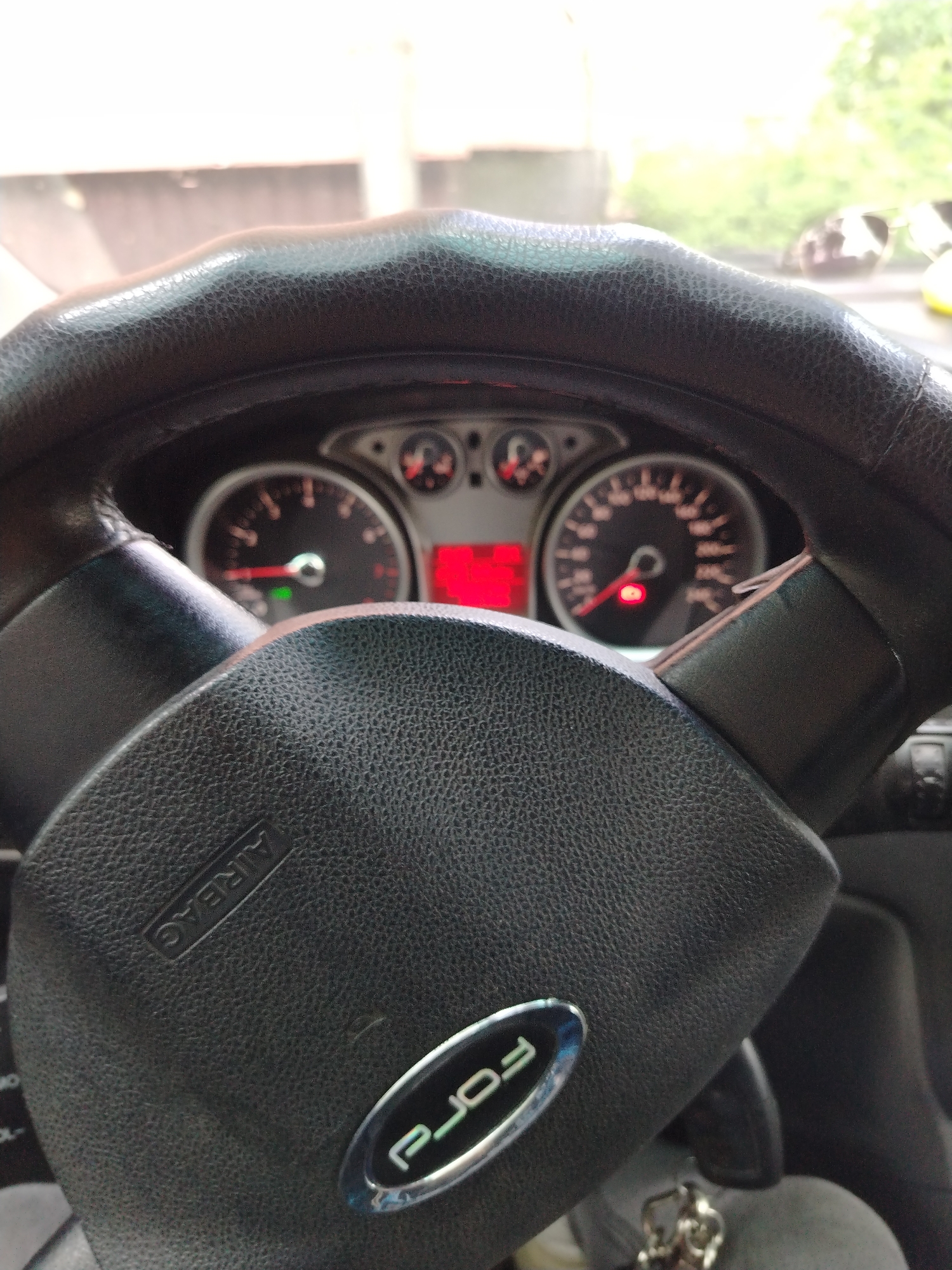 The image shows the interior of a car, specifically the steering wheel and dashboard. The steering wheel is black and has a "Ford" logo. The dashboard instruments are visible, including the speedometer and tachometer, with the car seemingly idling. The environment outside the car is blurred, but there appears to be greenery and possibly another vehicle with sunglasses on the dashboard. I cannot ascertain the location of Yaroslavl, Russia from the image.