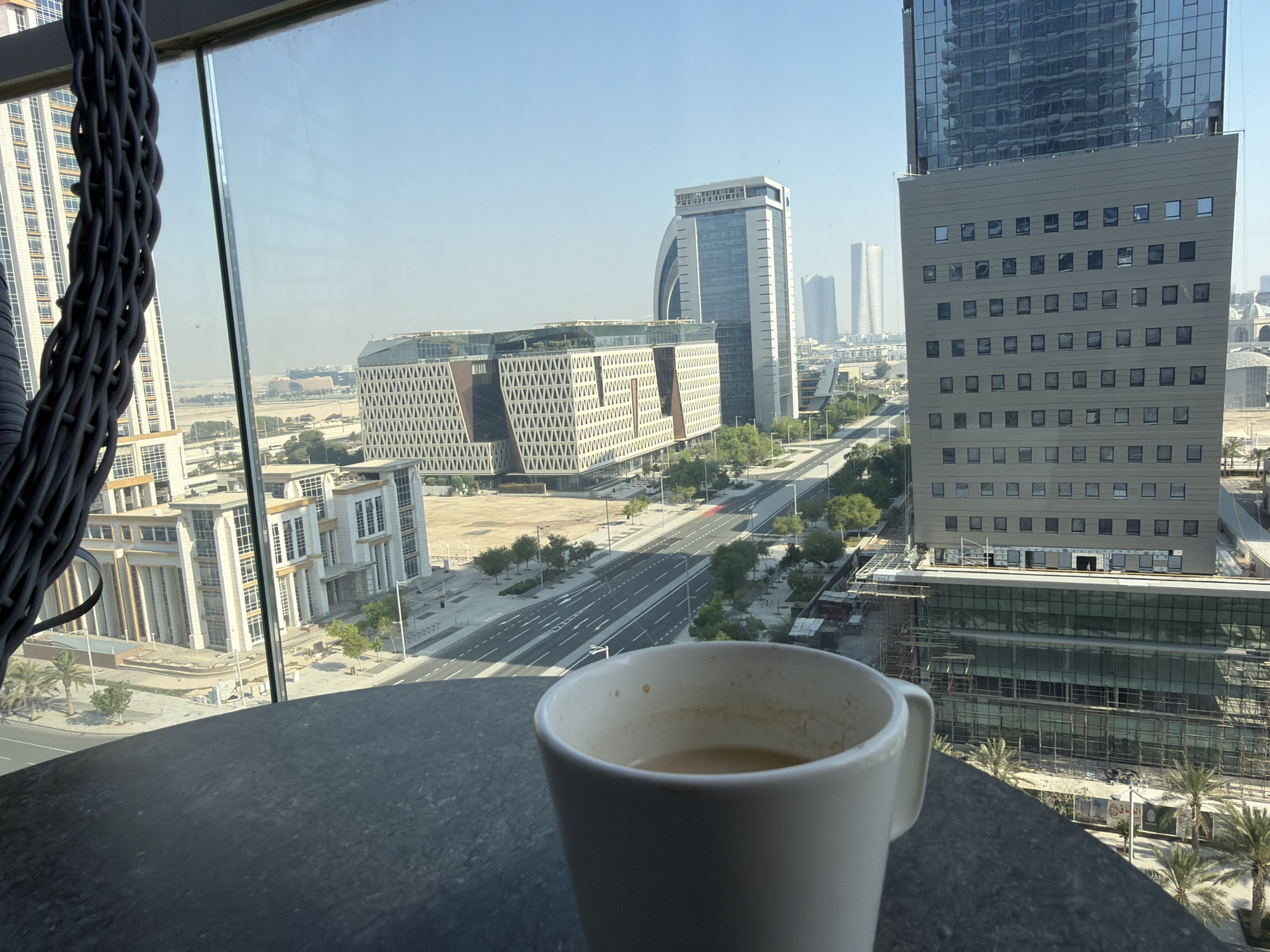 A close-up shot of a white mug with coffee sits on a dark, textured table in the foreground. Beyond the mug, a cityscape unfolds, viewed through a large window with a woven rattan chair partially visible on the left. The city is characterized by modern architecture, including several tall buildings and lower-rise structures with distinct geometric patterns. A wide, multi-lane road runs through the center of the scene, with sparse traffic. Palm trees and other greenery are interspersed throughout the urban landscape. The sky is clear and bright blue, suggesting a sunny day. The overall impression is one of urban sophistication and a moment of quiet contemplation, possibly from a high vantage point in a city.