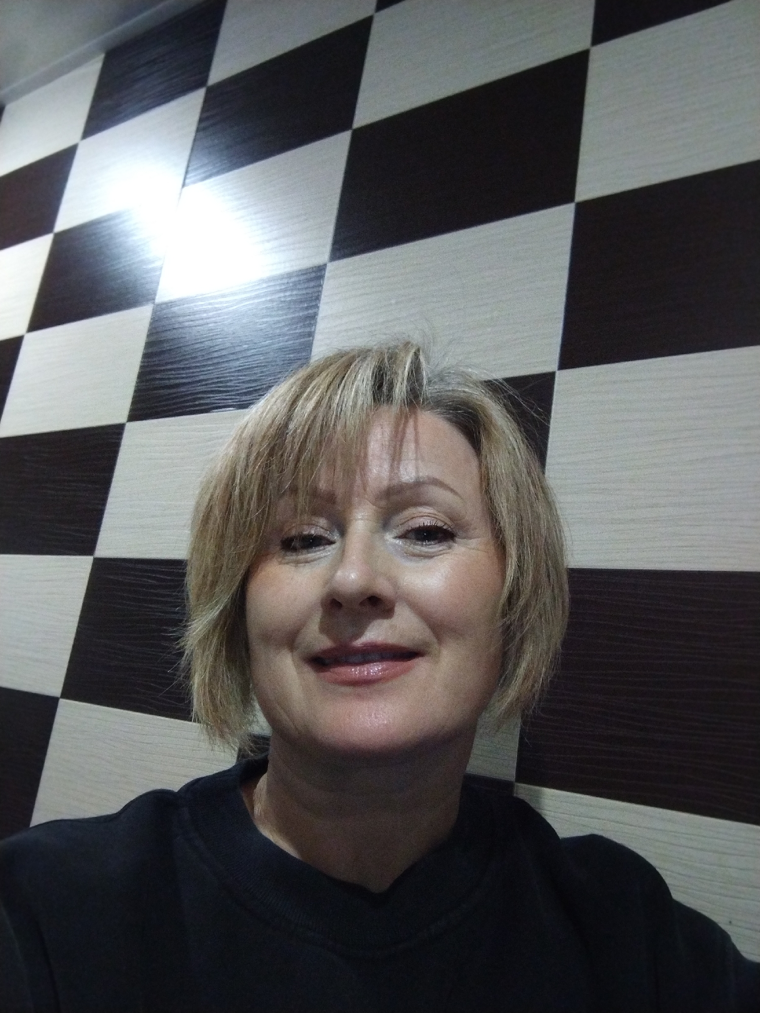 A medium shot photograph shows a Caucasian woman with short, blonde hair and fair skin, looking directly at the camera. She is wearing a black crew neck sweatshirt. Her face is partially illuminated by a light source positioned above and to the left of the frame, creating a sheen on her forehead and the bridge of her nose. Her eyes are open and looking forward. Her lips are slightly parted, revealing a subtle smile. The background consists of a wall with a checkerboard pattern of alternating light beige and dark brown rhombuses. The rhombuses have a wood grain texture. The lighting in the scene is moderately bright, with a distinct glare on the wall in the upper left quadrant. The photograph was taken in Bialynichy, Belarus.