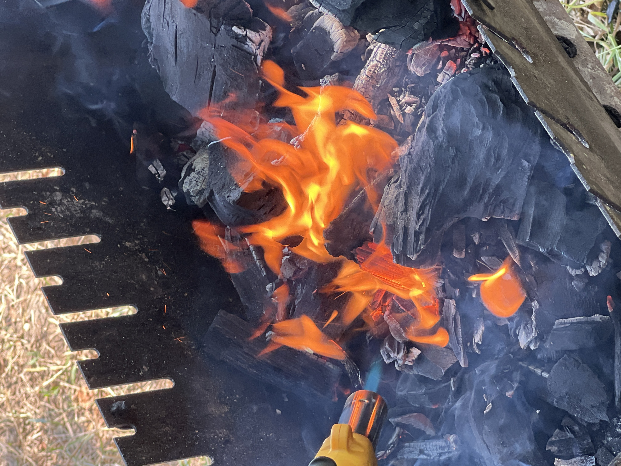 The image shows a close-up view of a barbecue grill filled with charcoal and flames. A yellow butane torch is being used to ignite the charcoal on the left side of the frame, producing a blue flame. The grill itself appears to be made of metal and has a serrated edge along the top. Wisps of smoke rise from the burning coals. The background is blurred, showing dry grass and dirt, suggesting an outdoor setting. The scene is consistent with preparing a fire for grilling. No specific location within Ждановіцкі сельскі Савет is identifiable from the image.