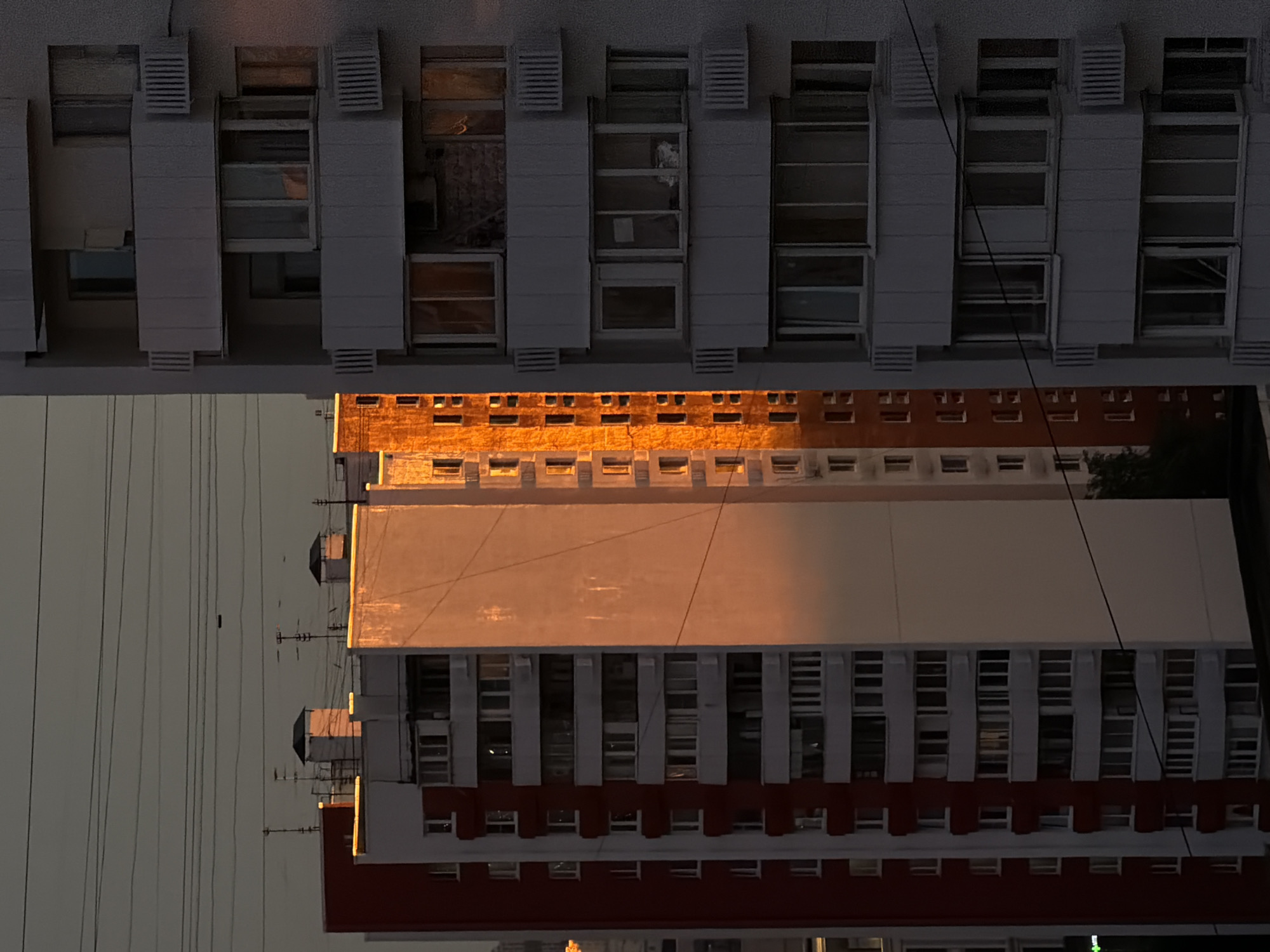 The image captures an exterior view of several apartment buildings in Khimki, Russia, oriented vertically. The primary subjects are the architectural structures, characterized by their repetitive window patterns and light-colored facades. The time of day appears to be sunset or sunrise, as indicated by the warm, golden light illuminating parts of the buildings. The sky is a muted, overcast gray, suggesting a cloudy or hazy atmosphere.
The scene is urban, with visible power lines crisscrossing the sky, hinting at the presence of electrical infrastructure common in residential areas. The buildings themselves feature balconies, air conditioning units, and brickwork accents. One section of a building has a distinct orange-red color band running horizontally, contrasting with the lighter upper levels. Another building's upper section is bathed in a bright, warm light, highlighting its textured surface. The lower portions of the buildings are in shadow, and the windows reflect the sky and surrounding structures.
There are no people or specific activities visibly depicted. The focus is on the static architecture and the play of light on the building surfaces. The composition, with its vertical orientation and the stark lines of the buildings and wires, creates a somewhat impersonal and geometric feel to the urban landscape. No text is discernible in the image.