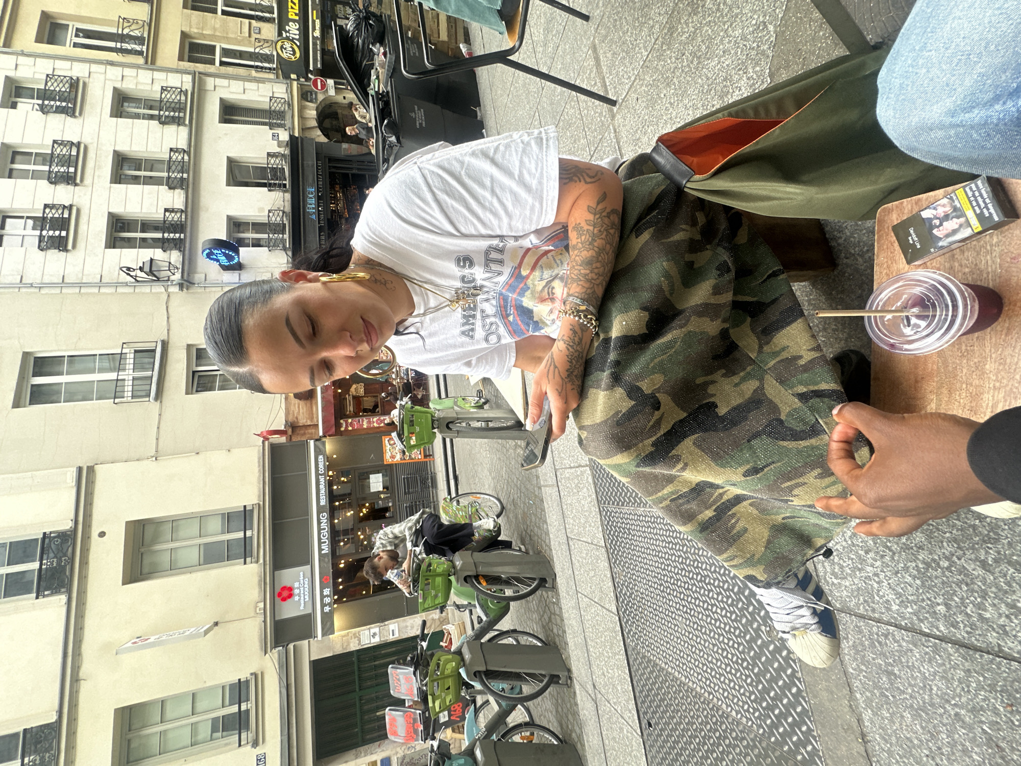 The image shows a woman with tattoos sitting at an outdoor cafe in Paris, France. She is wearing a white t-shirt with an "America's Most Wanted" graphic and camouflage pants. She has gold hoop earrings and is holding a phone in her hands. Next to her on the table is a drink with a straw and a pack of Davidoff Line cigarettes.
In the background, there is a building with apartments and shops on the ground floor. There are several bicycles parked nearby. The street is paved with gray stones. A sign for "Mugung Restaurant Coréen" is visible.