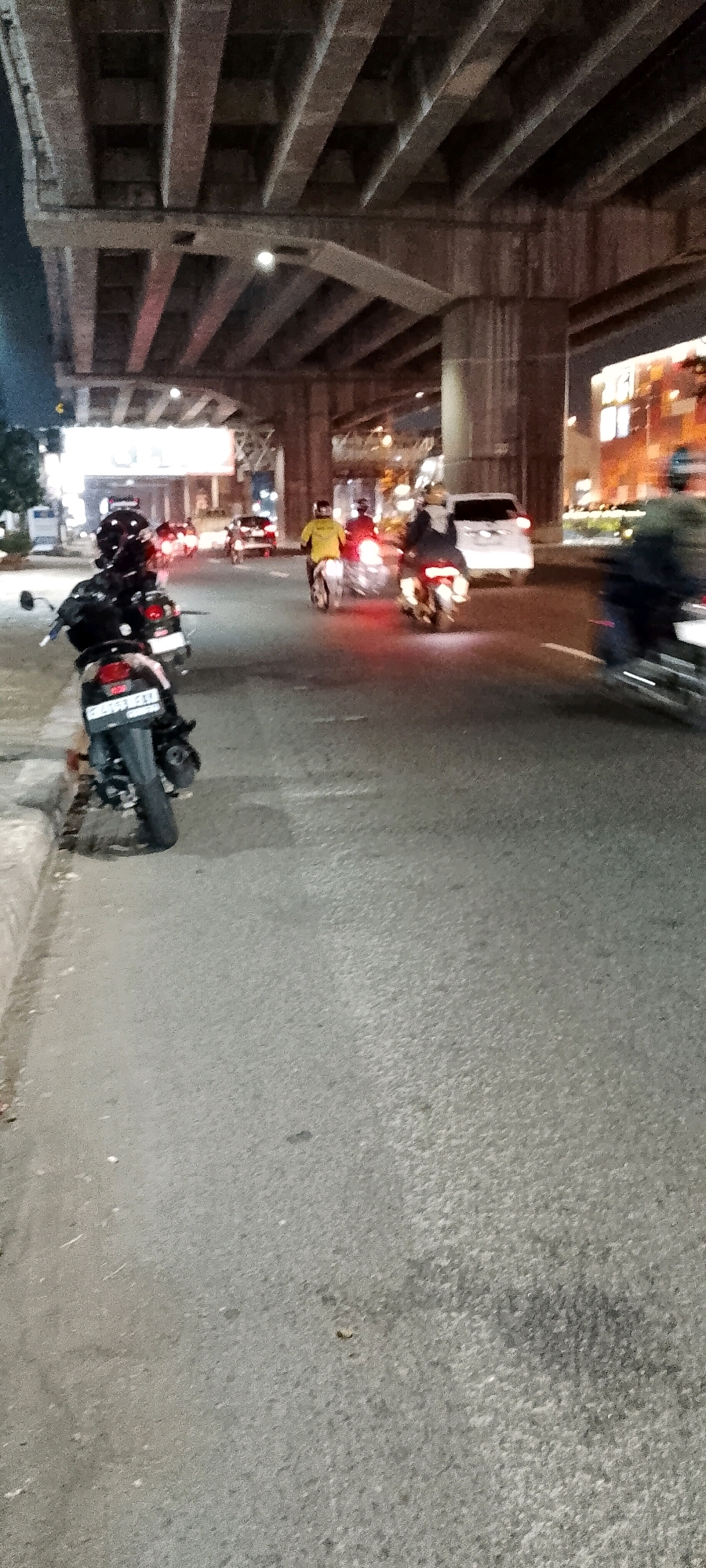 This is a ground-level, vertical, slightly blurry, nighttime photograph of a busy street in Bekasi, Indonesia, underneath an elevated structure. The scene is dominated by a concrete overpass with multiple beams and supports. The road is asphalt, with a concrete curb on the left. Several motorcycles are visible on the road. In the foreground left, a black motorcycle is parked, facing right, with its license plate visible. Further down the road, other motorcycles, some with riders wearing helmets and jackets, are in motion, indicated by motion blur. One rider wears a yellow jacket. On the right side of the road, headlights and taillights of vehicles are visible. Buildings with illuminated windows are present in the background on the right, suggesting an urban environment. The overall impression is a lively, active nighttime street scene in Bekasi.