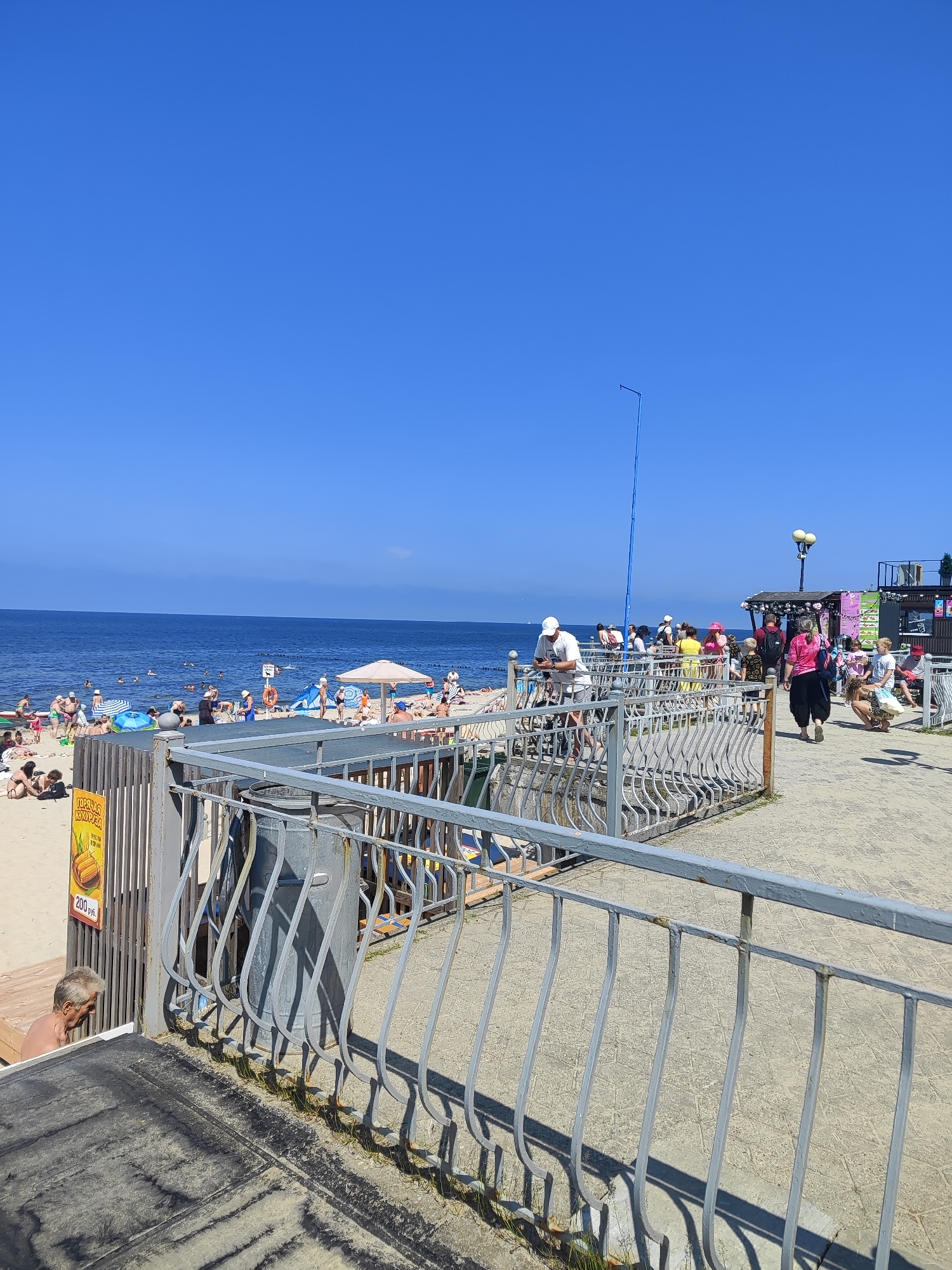 The image depicts a sunny day at a beach in Pionersky, Russia. The foreground features a grey metal railing and a dark surface covered in sand. Behind the railing, a paved walkway is populated by a diverse group of people. Some are strolling, while others are gathered in small groups. A few individuals are seen in the background at a food stall or cafe. To the left of the walkway, a sandy beach is visible with numerous people sunbathing and swimming in the blue ocean. Beach umbrellas provide shade on the sand. The sky is clear and a vibrant blue.