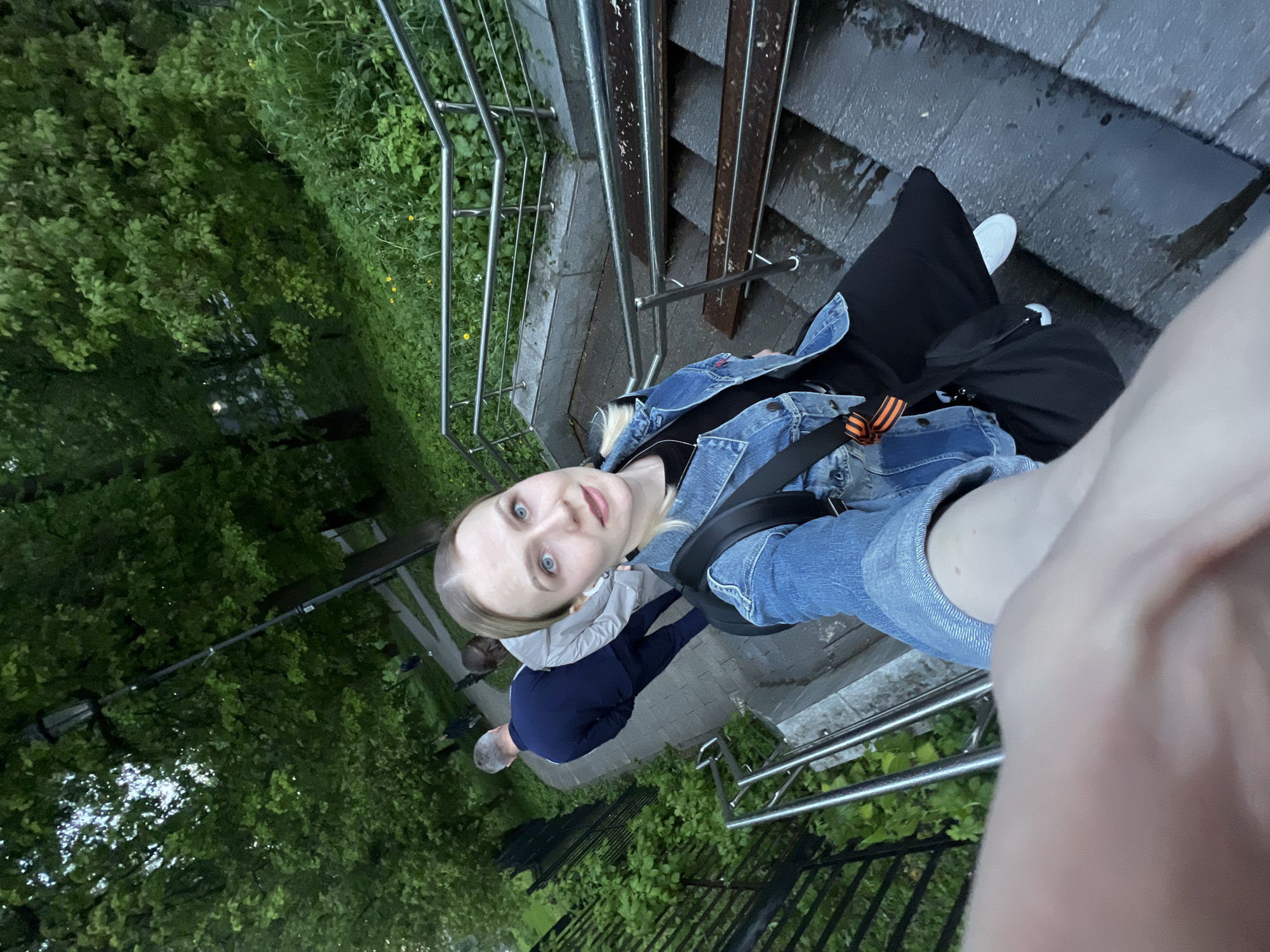 A young woman is taking a selfie on a staircase. She is wearing a denim jacket, a black shirt, and black pants. She has a black backpack on her shoulder. She is standing on wet stone steps with metal railings on both sides. In the background, there is a park with green trees. Two people are standing behind her, seemingly walking on a pavement. The scene appears to be in a city, specifically Khimki, Russia.