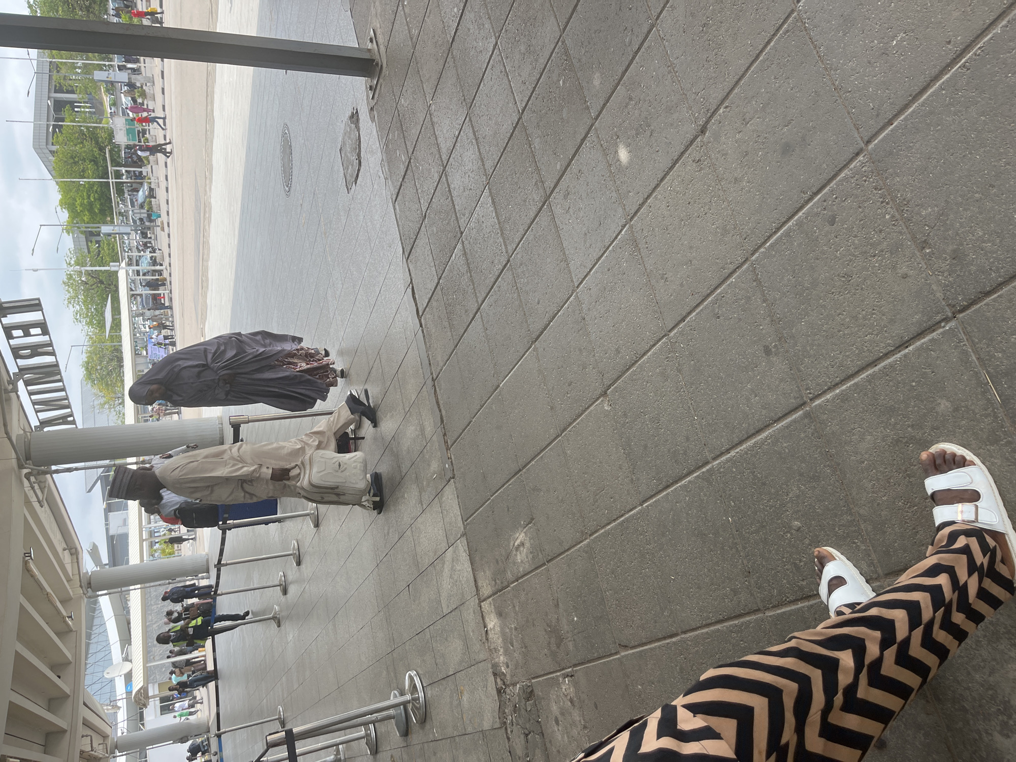 The image captures a vibrant daytime scene at what appears to be the exterior of an airport or a large public transportation terminal. The camera is positioned low, looking outwards from beneath a building structure towards an open area and a road.
In the immediate foreground, the legs and feet of a person are prominent. They are wearing wide-legged pants with a distinctive black and beige/light brown chevron pattern and white flat sandals with two buckles, the left foot slightly elevated. The ground is paved with large, rectangular dark grey tiles, some showing signs of wear.
In the mid-ground, two individuals are walking away from the camera. To the left, a person is dressed in light-colored traditional attire, possibly an agbada with a matching cap, carrying a light-colored shoulder bag. To their right, another person wears a dark, long, flowing garment, perhaps an abaya, with a patterned skirt visible underneath.
The background reveals a bustling environment. Multiple people are visible, some waiting in lines behind silver stanchions, suggesting an organized queue or security checkpoint. Others are walking freely. Further back, a wide paved area serves as a road or parking lot, populated with various vehicles, including cars and buses. Modern-looking buildings with large pillars, trees, and streetlights frame the distant horizon under an overcast sky.
A significant detail is the visible text "TERMINAL," written vertically on a sign or building structure in the far left of the background, confirming the location as a terminal building. The overall lighting suggests it is daytime with diffused light, possibly due to an overcast sky. The activity depicted points to routine movement and waiting typical of a busy transport hub. Given the context, this scene is set in Abuja, Nigeria.