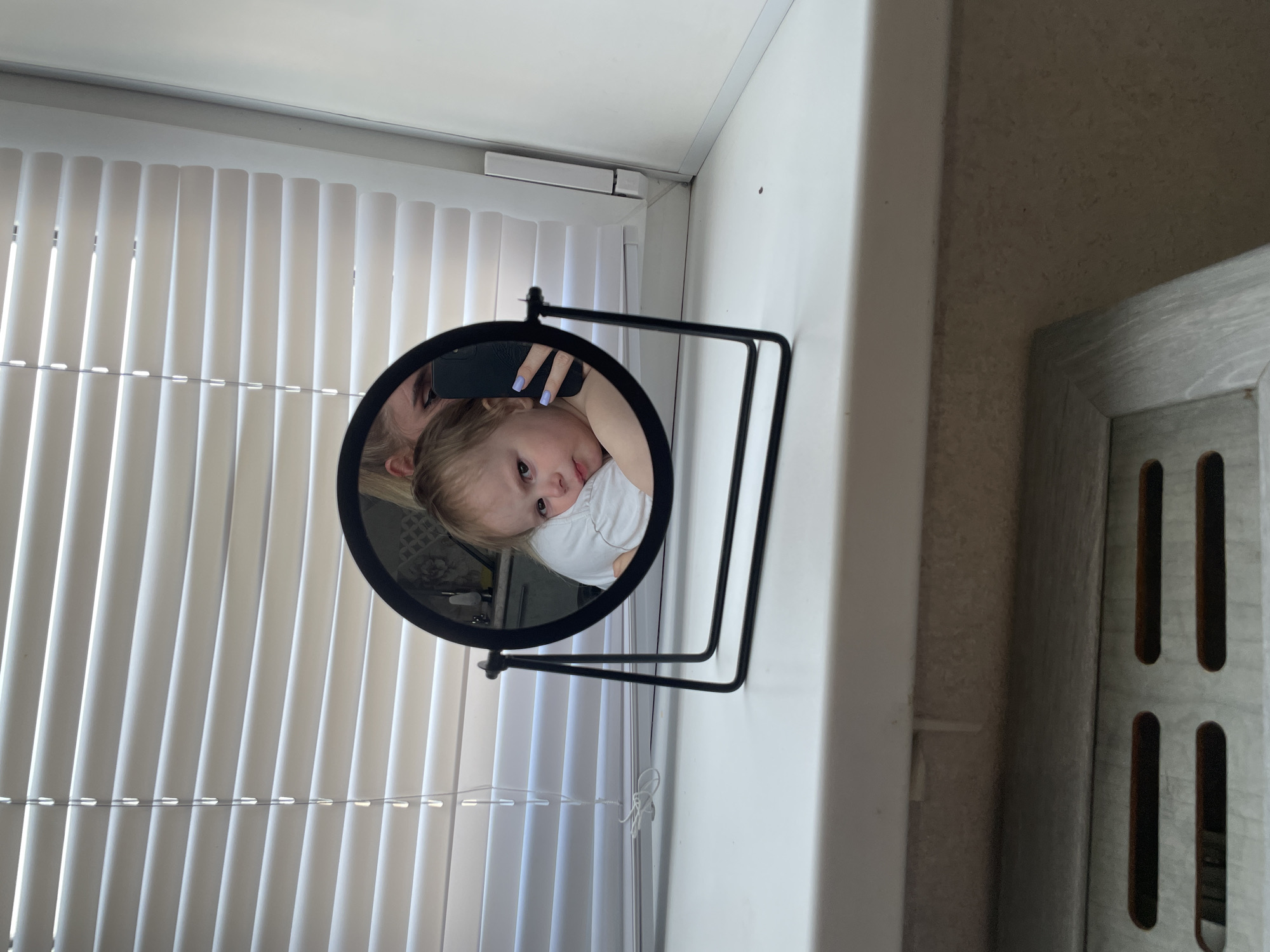 The image captures a reflection in a round tabletop mirror, which is positioned on a white window sill in an indoor setting. Inside the mirror, a young child with light hair and a white t-shirt is visible, looking calmly towards the viewer. Behind the child's head, an adult's hand holding a dark-cased smartphone is reflected, indicating the adult is taking the photograph. The adult's fingernails are painted a light purple or lavender.
The mirror has a black frame and a black metal stand. In the background of the actual room, vertical white blinds cover a window, allowing daylight to filter through. A thin cord for the blinds is visible near the base of the mirror's stand. Above the blinds, a small white rectangular object is mounted on the wall or window frame. The walls visible in the scene are white. To the right of the window sill, a textured wall section and a light-colored wooden frame with vertical slots (possibly part of a vent or furniture) are partially visible. No text is discernible within the image. The scene appears to be taking place during the daytime. While the context suggests Vladivostok, Russia, there are no specific visual cues within the image to confirm this location.