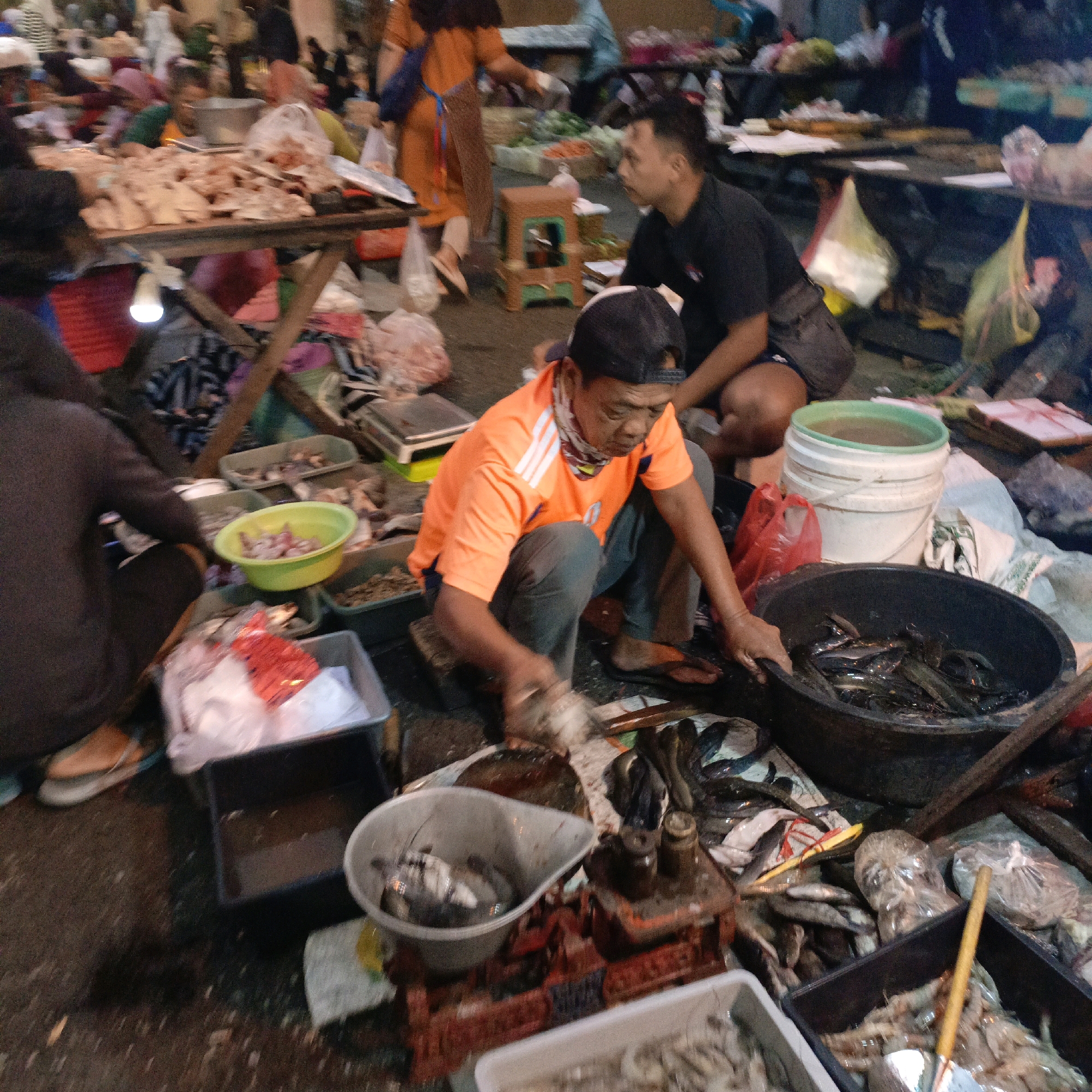 The image depicts a bustling outdoor market scene, likely in Salatiga, Indonesia, given the context. In the foreground, a man wearing an orange t-shirt and a black cap is squatting and appears to be handling fish, possibly preparing them for sale. He is surrounded by various containers and displays of seafood, including a large black tub filled with fish, and a green bowl containing smaller items. To his left, a person wearing a dark hooded jacket is also seated, facing away from the camera. Further back, a woman in an orange dress and headscarf is visible, tending to a stall with what appears to be raw meat. Other individuals and market stalls are present in the background, creating a sense of lively commerce. The ground is wet and appears to be unpaved, typical of many traditional Indonesian markets. The overall atmosphere is that of a busy marketplace with vendors and customers engaged in transactions.