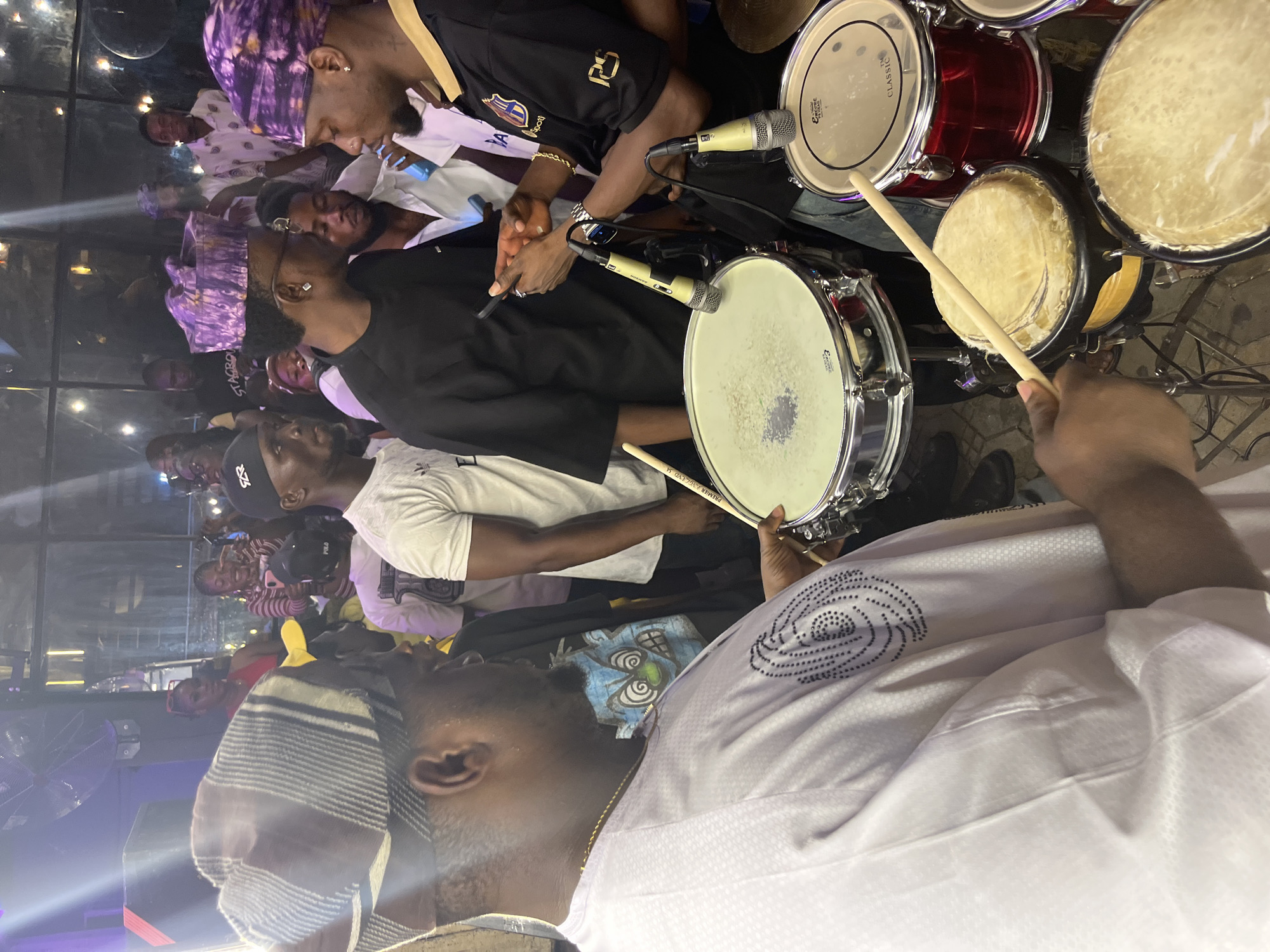 In an indoor setting, possibly a nightclub or concert venue, a group of people are gathered, with a focus on a drum set. The foreground is dominated by a man wearing a light-colored, patterned traditional outfit and a striped head covering, holding a drumstick and positioned as if playing a drum. To his right, another man in a black shirt and a purple patterned head covering is visible, looking towards the first man. In the mid-ground, several other individuals are present, some wearing similar head coverings. The background reveals a dimly lit environment with a stage area, stage lighting, and a crowd of onlookers. The drum set includes multiple drums and microphones, suggesting a musical performance is underway or about to commence. The overall atmosphere appears lively and celebratory. The city is unknown, as is the specific location within the city.