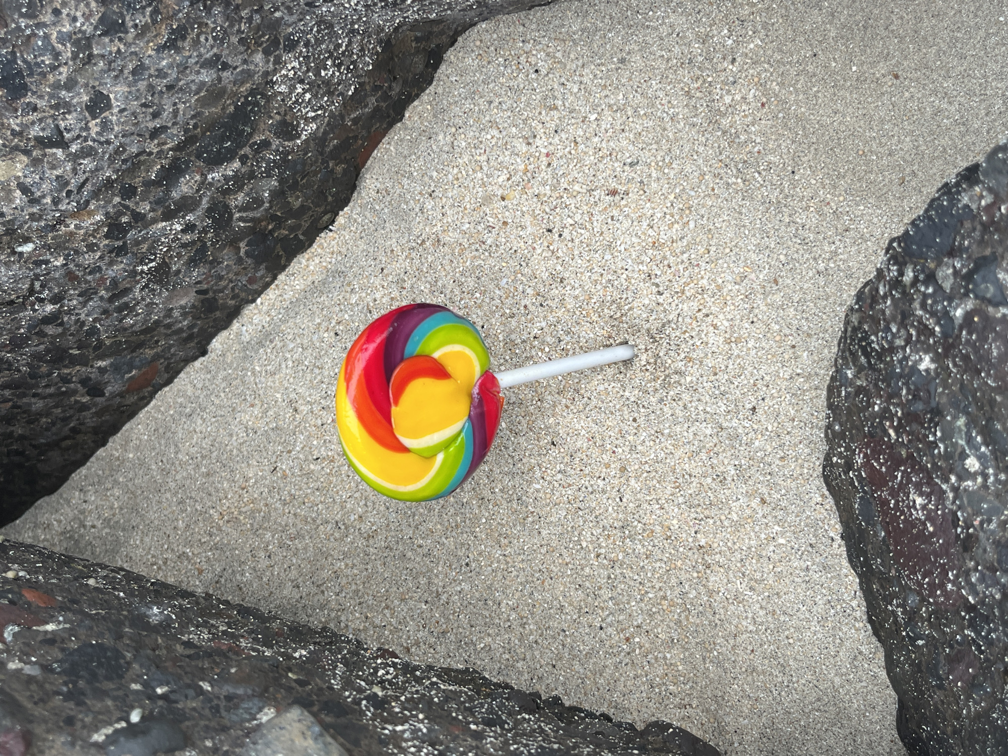 A rainbow swirl lollipop rests on fine-grained sand, nestled between dark, coarse-textured rocks. The lollipop, positioned slightly off-center to the right, features vibrant bands of red, orange, yellow, green, blue, and purple. Its white stick is partially embedded in the sand. The sand itself is a light beige color with visible small shell fragments and pebbles. The rocks form a triangular enclosure, suggesting a natural crevice or a man-made structure, possibly a breakwater or part of a coastal defense system. The lighting suggests it is either dusk or dawn, or an overcast day, as there are no harsh shadows. The scene appears to be a secluded spot along the coast, a detail that would be consistent with many of Denpasar's beaches, such as Seminyak Beach or Sanur Beach, which feature similar rocky outcrops and sandy shores.