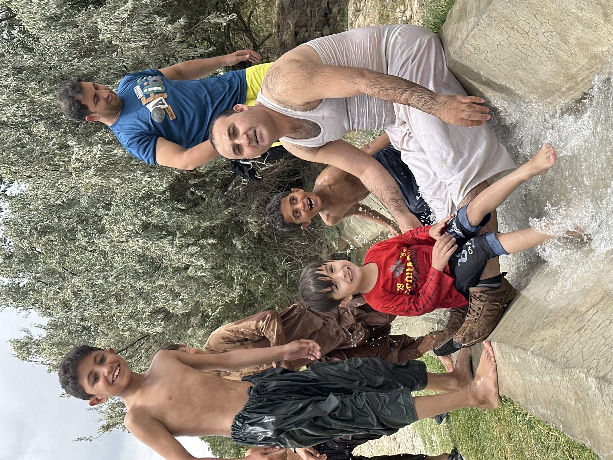In Kan Mehtarzai, Pakistan, a group of males, including at least three children and two adults, are seen in what appears to be an outdoor water channel or ditch. The environment suggests a natural setting with a large tree with light-colored foliage dominating the background. The main subjects are positioned in and around the water channel.
In the foreground, a young boy in a red long-sleeved shirt and dark pants is seated, with water splashing around his legs. He is smiling and looking towards the camera. To his left, a young boy with no shirt on and dark, baggy pants stands with a wide, open-mouthed smile, suggesting excitement. Another adult male, wearing a white tank top and light-colored, loose-fitting pants, is seated in the water channel to the right of the children, looking directly at the camera.
Behind these individuals, another young boy, shirtless, is visible. Further back, an adult male in a blue t-shirt and yellow shorts stands, looking down. Another adult in a brown, traditional garment stands partially obscured by the others. The water in the channel is actively flowing, creating splashes and ripples around the individuals. The ground around the channel appears to be covered in grass.