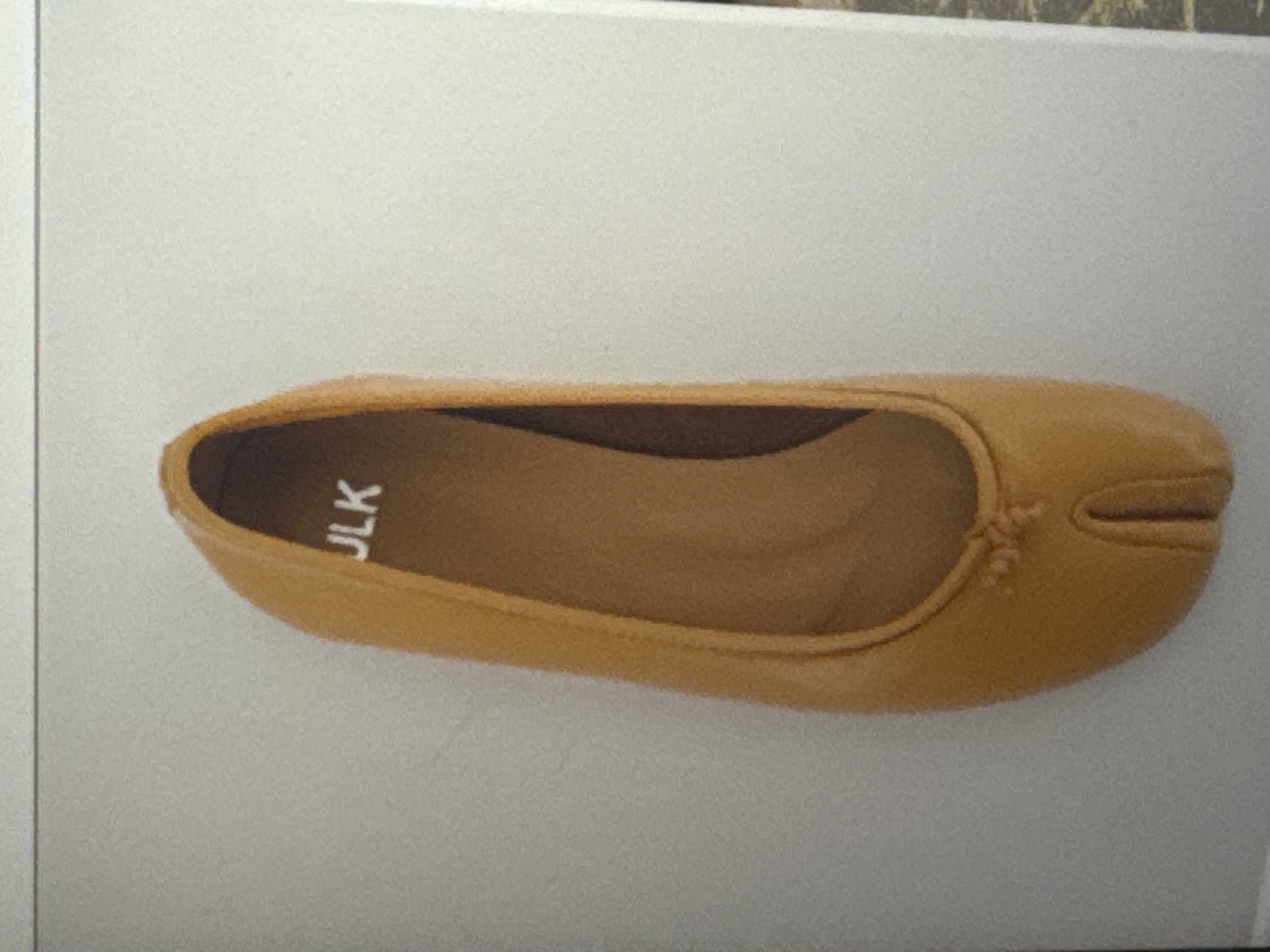 A single, tan-colored Tabi-style ballet flat is displayed against a pale grey, textured surface. The shoe is positioned vertically with the toe pointing upwards. The interior of the shoe is a darker tan color and bears the white imprinted text "JLK". The shoe has a distinctive split toe design. The background appears to be a wall or a large flat surface. There is no discernible connection to Tbilisi, Georgia.