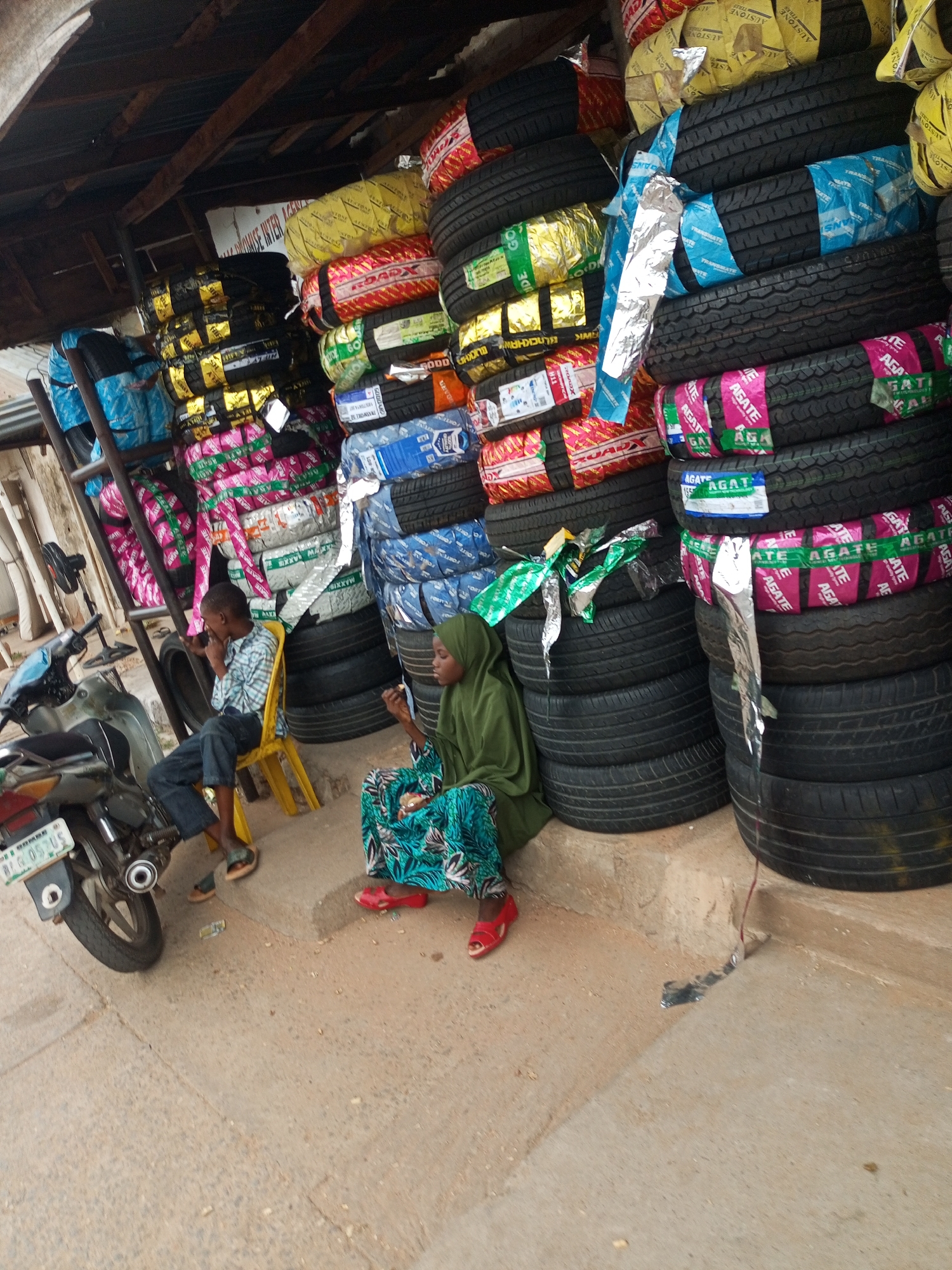 Arawa, Nigeria. This is a street-level view of a tire shop, packed with various types of vehicle tires stacked high. The shop appears to be open-air, with a corrugated metal roof supported by wooden beams. In the foreground, a young boy, dressed in a checkered shirt and dark trousers, sits on a bright yellow plastic chair. To his right, a woman wearing a green hijab and a blue and green patterned dress is seated on the ground, holding what appears to be food. A motorcycle is parked to the left of the boy. The ground is dusty and uneven, suggesting a rural or developing urban environment. The tires are wrapped in colorful packaging, some with prominent branding like "AGATE" and "ROADX". The overall impression is one of a busy commercial establishment in a hot, dusty climate.