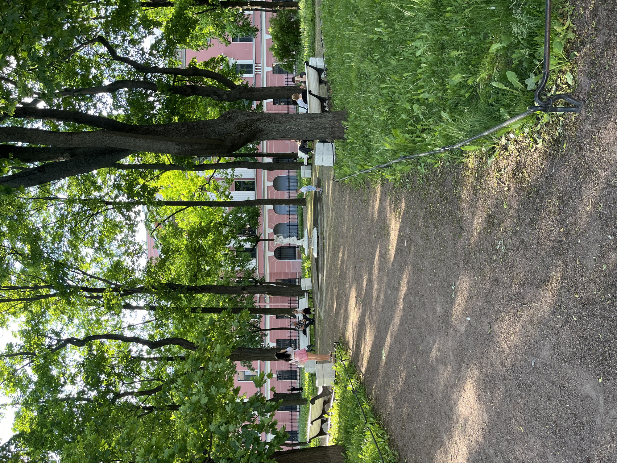 The image depicts a sunlit park scene, identified as the Anichkov Garden in Saint Petersburg, Russia, with the distinct pink facade of the Anichkov Palace visible in the background.
In the foreground, a wide, light brown dirt or gravel path traverses the scene, marked by dappled sunlight and shadows from overhead trees. To the right of the path, a low, dark metal fence separates it from an area of lush green grass with various plants and weeds. Two unoccupied park benches with white slats and dark metal frames are positioned along the left side of the path.
In the midground, a young woman with dark hair, wearing a white short-sleeved top and a bright pink pleated mini-skirt, stands facing right, near the path. Her arms are down, and she appears to be holding an object. Further into the park, a small white decorative fountain or sculpture, possibly featuring a figure, is centrally located within a circular paved area. A small child, dressed in light blue shorts and a white top, is visible walking near the fountain towards the center. Several other individuals are seated on white-slatted park benches positioned beyond the fountain: two people on a bench to the left and at least three people on a bench to the right, some appearing to be engaged in conversation. Numerous mature trees with dark, thick trunks and abundant green foliage are scattered throughout the park, providing shade and casting shadows. Sunlight filters brightly through their leaves.
The background is dominated by a grand, multi-story historical building featuring a prominent light pink facade with dark rectangular windows and white architectural accents, including arched recesses on the ground floor. This building is identified as the Anichkov Palace. A black, ornate metal fence stands in front of the palace, separating it from the park area. The upper portions of the palace are partially obscured by the dense tree canopy. The sky, glimpsed through the leaves, appears bright and clear. The overall lighting suggests a sunny day.
