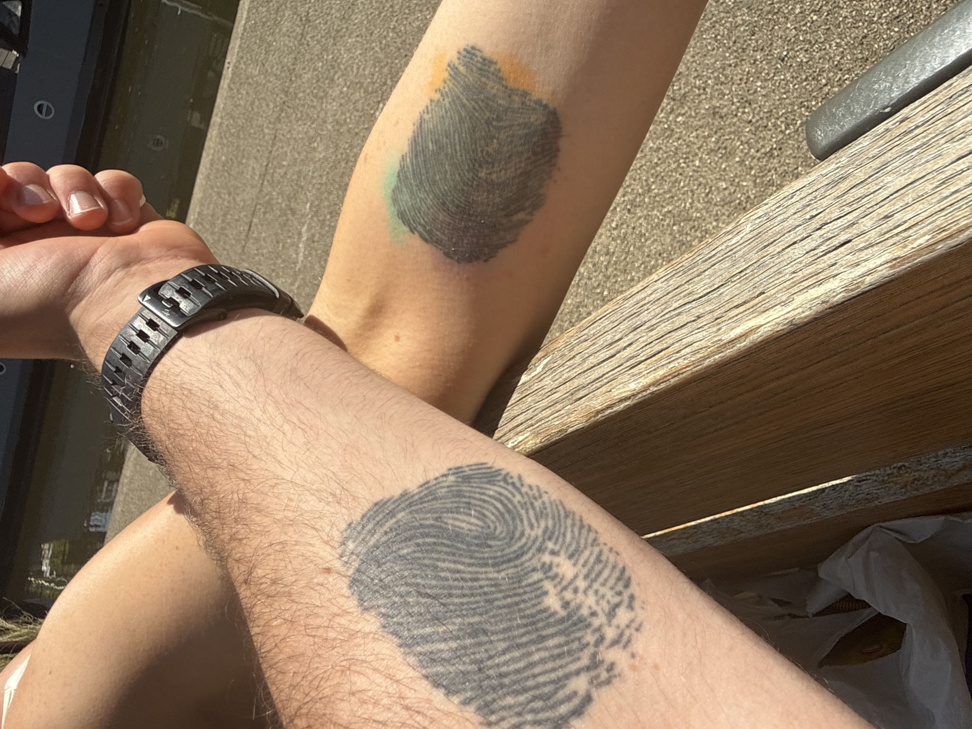 The image shows two arms with fingerprint tattoos. The left arm wears a black watch. The location appears to be outdoors, possibly a park or public area, based on the presence of a wooden bench and water visible in the background. The city and state are unknown.