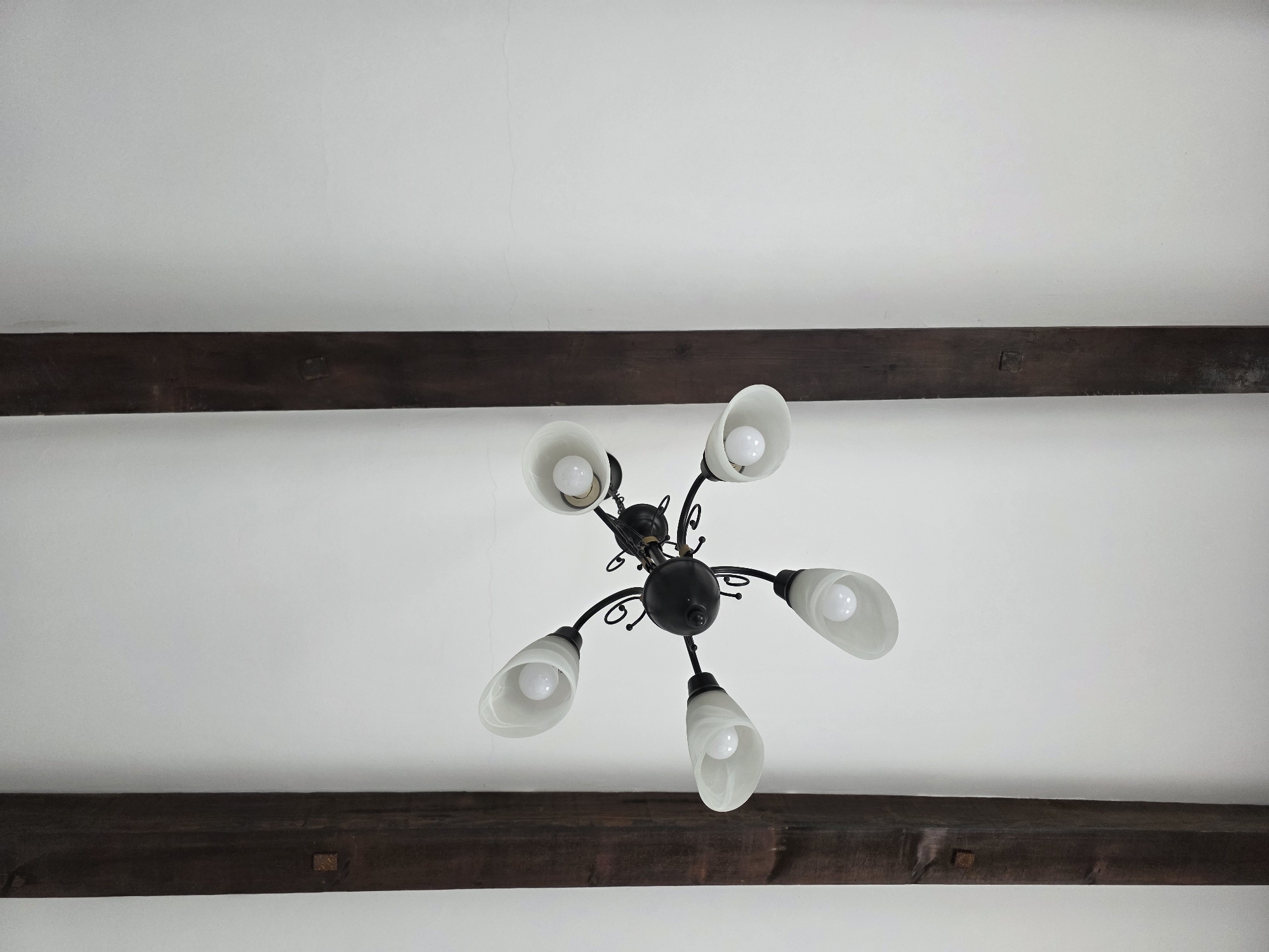 The image depicts a ceiling with exposed wooden beams and a central chandelier. The beams are dark brown and run horizontally across the frame. The chandelier is black with five upward-facing white glass shades, each containing a light bulb. The setting appears to be an interior space in Ulcinj-Ulqin, Montenegro, though precise localization is not possible without more information.