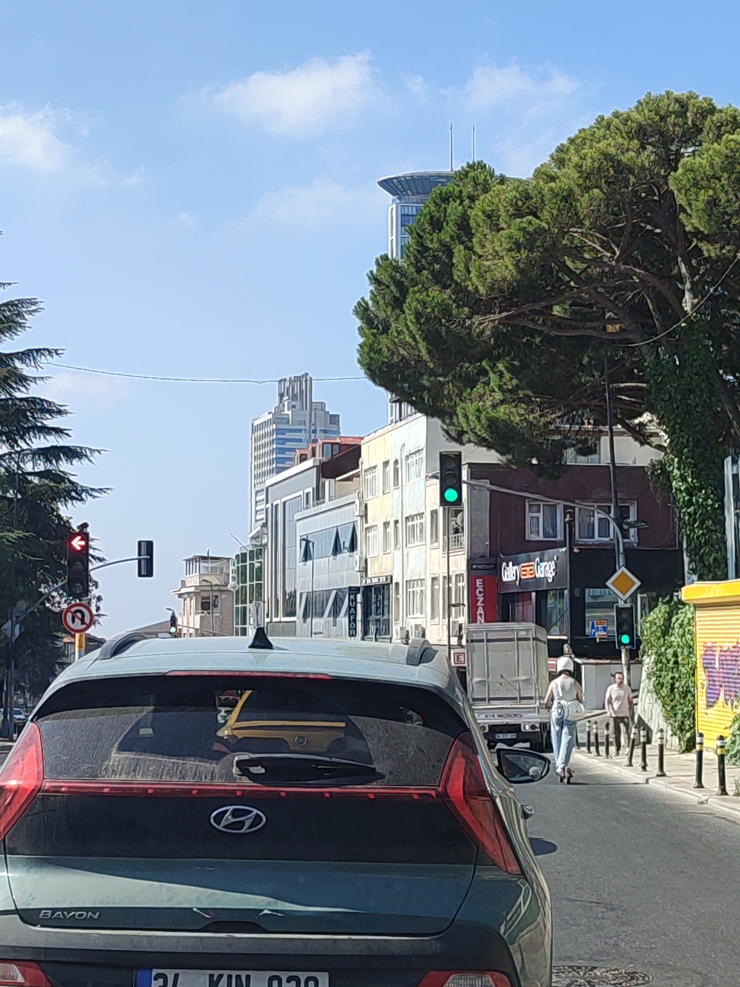 In Üsküdar, Turkey, on a clear, sunny day, a street scene unfolds with a focus on a busy intersection. The foreground is dominated by the rear of a dark green Hyundai Bayon car, its taillights and distinctive logo visible. Beyond this vehicle, the road continues, lined with buildings of varying architectural styles, some modern and glass-fronted, others older with more traditional features. Lush green trees, including a large pine tree, frame the scene, their branches extending over the street and sidewalks.
Traffic lights play a significant role in the composition, with one showing a red signal and an arrow pointing left, indicating a turn restriction, while another further down the road displays a green light. A traffic sign indicating "no U-turn" is also present. People are visible in the mid-ground: a person riding an electric scooter, wearing a white helmet and light blue jeans, heads away from the viewer. Behind them, a man in a light-colored t-shirt and dark pants stands on the sidewalk. Further back, a white truck with "KIA MOTORS" branding is stopped at the intersection. Several bollards line the sidewalk, and a yellow structure, possibly a temporary barrier or construction element, is seen on the right side of the image. The sky is bright blue with scattered white clouds. The scene suggests a typical urban environment in a city undergoing development, with a mix of old and new infrastructure.