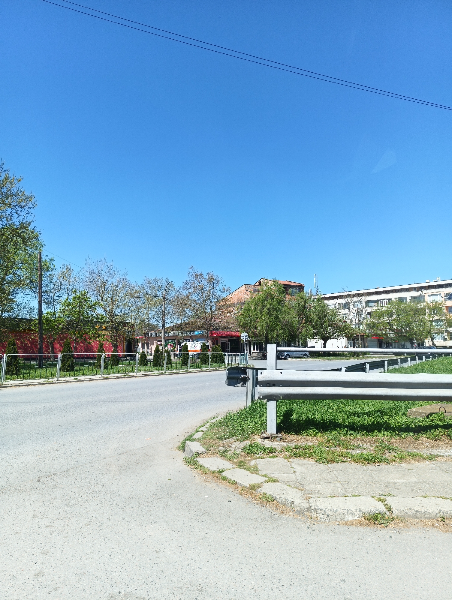 The image shows an urban street scene in Straldzha, Bulgaria, on a sunny day. The sky is clear and blue. The street pavement is light grey. A metal guardrail runs along a grassy area to the right of the street. Across the street, there's a red building with star decorations on its facade, partially obscured by trees. Further down the street, other buildings are visible, including a white building with multiple windows. A car is parked near the white building. Electrical wires stretch across the sky.