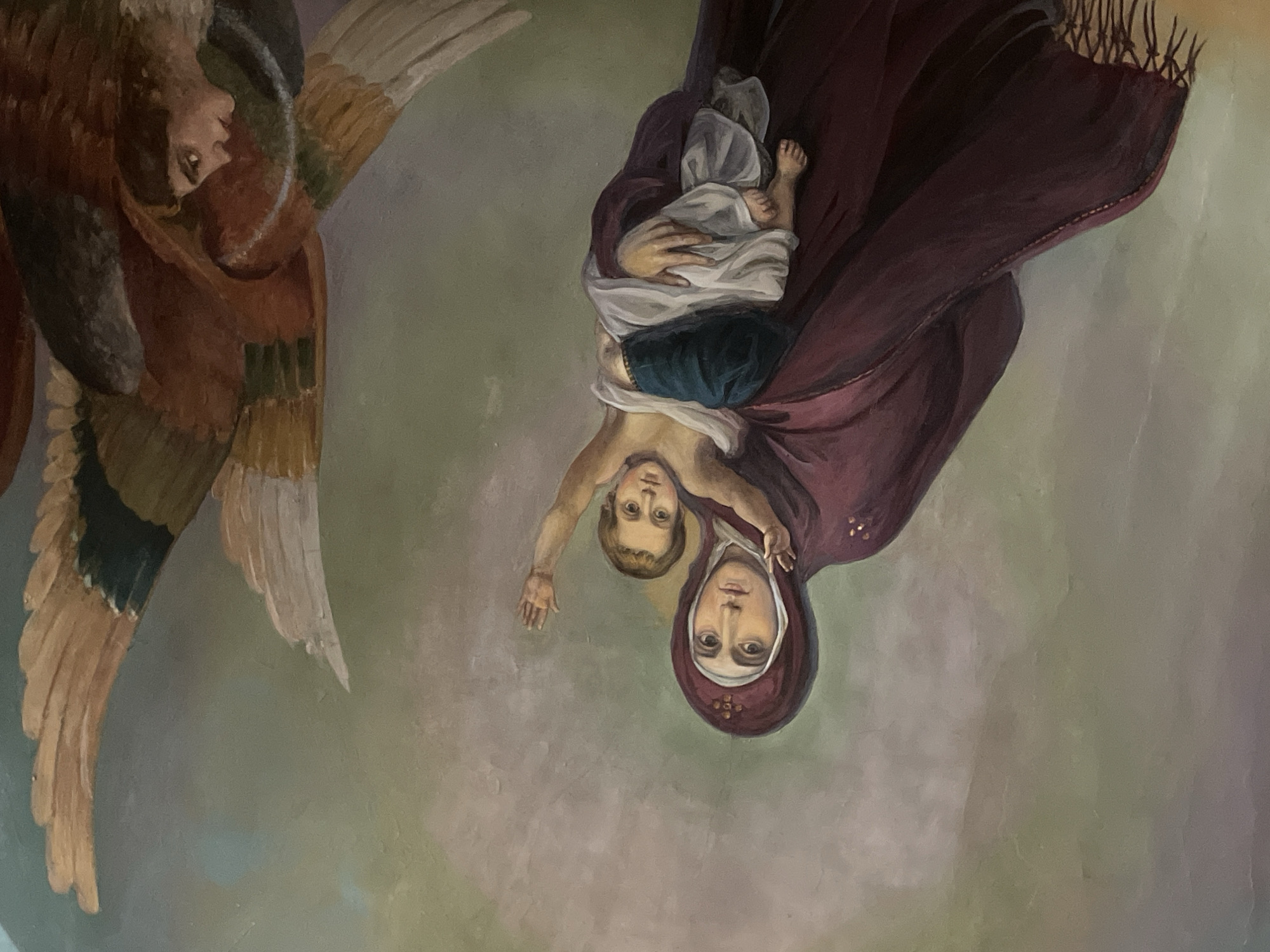 The image depicts a ceiling fresco in Moscow, Russia. It features a woman in a burgundy cloak and a white head covering with a red embellishment, holding a baby. The baby is wearing blue pants and a white garment. An angel with brown and white wings is positioned on the left side of the image. The background is a mottled blend of pale green and grey.