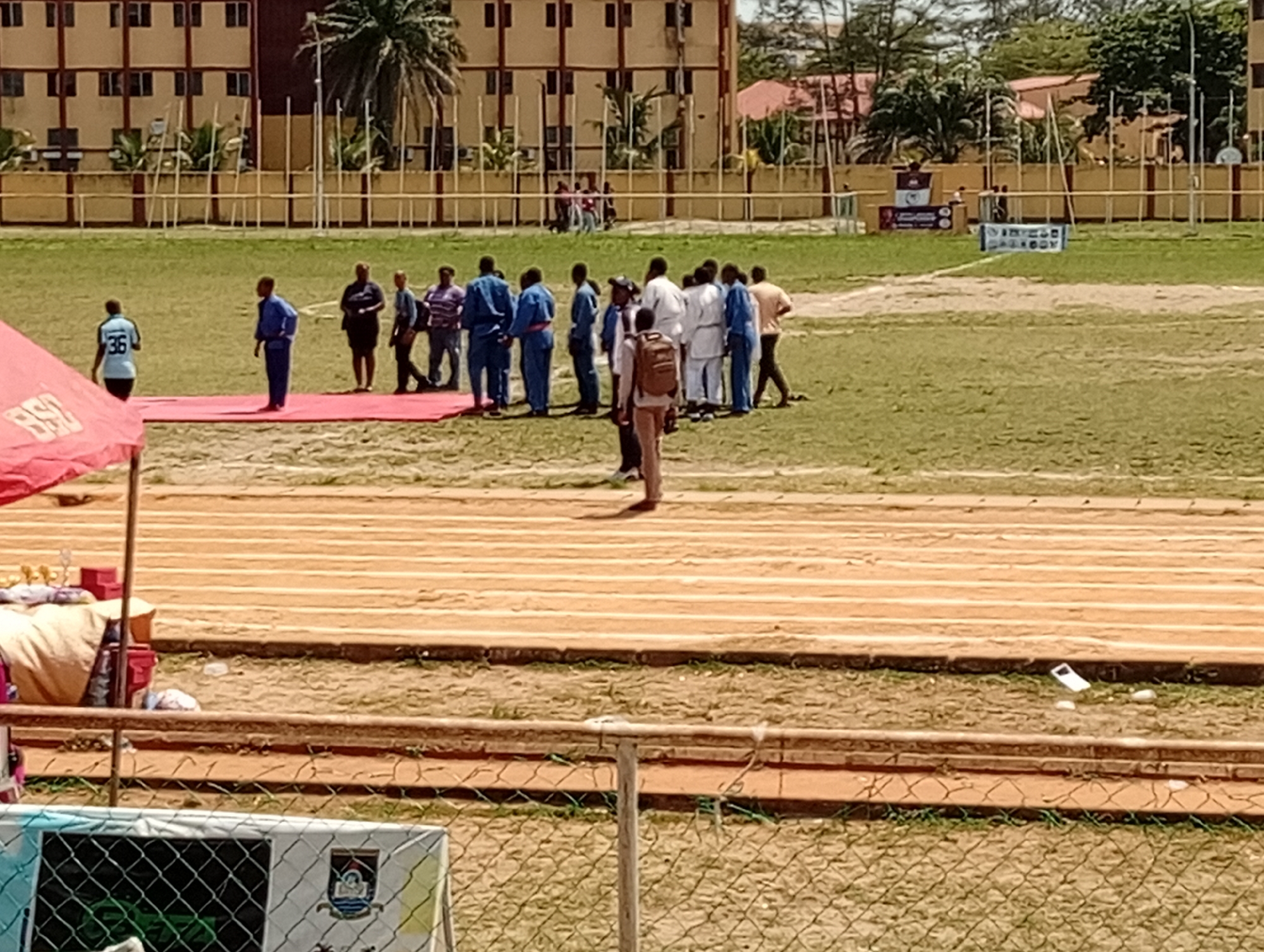 The image shows a sports field in an urban location in the city of Unknown, Unknown. A group of people, many wearing blue and white judogi, are gathered on the green field. A red mat is laid out nearby. In the foreground, there is a running track. A partial view of a fence is visible. In the background, there is a large building with multiple stories.