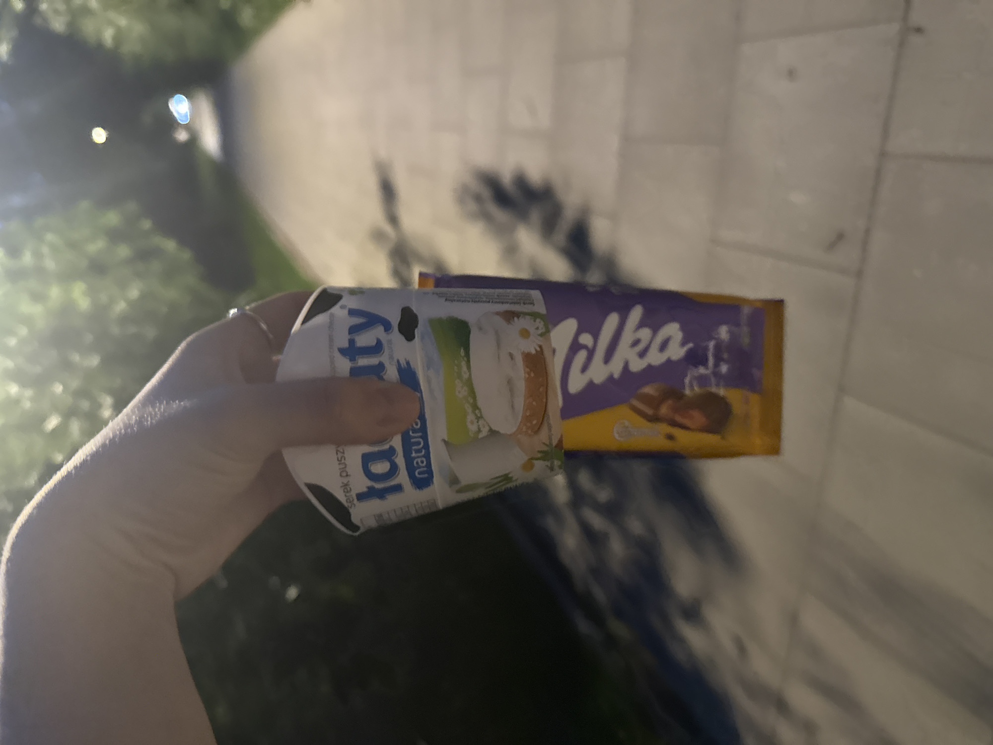 A hand, with a ring on the index finger, is holding a white and blue cup labeled "Łaciate" and a purple and yellow candy bar. The cup appears to contain a white, creamy substance and has an image of a pastry with a daisy on it. The candy bar's wrapper has the brand name "Milka" in large, purple lettering. The items are positioned in front of a paved walkway lined with trees, suggesting an outdoor, possibly urban, setting. The lighting indicates it is either dusk or nighttime, with some artificial lights visible in the background. The ground is paved with light-colored rectangular stones. Shadows are cast on the ground from the surrounding trees and vegetation. The overall scene is captured from a low-angle perspective.