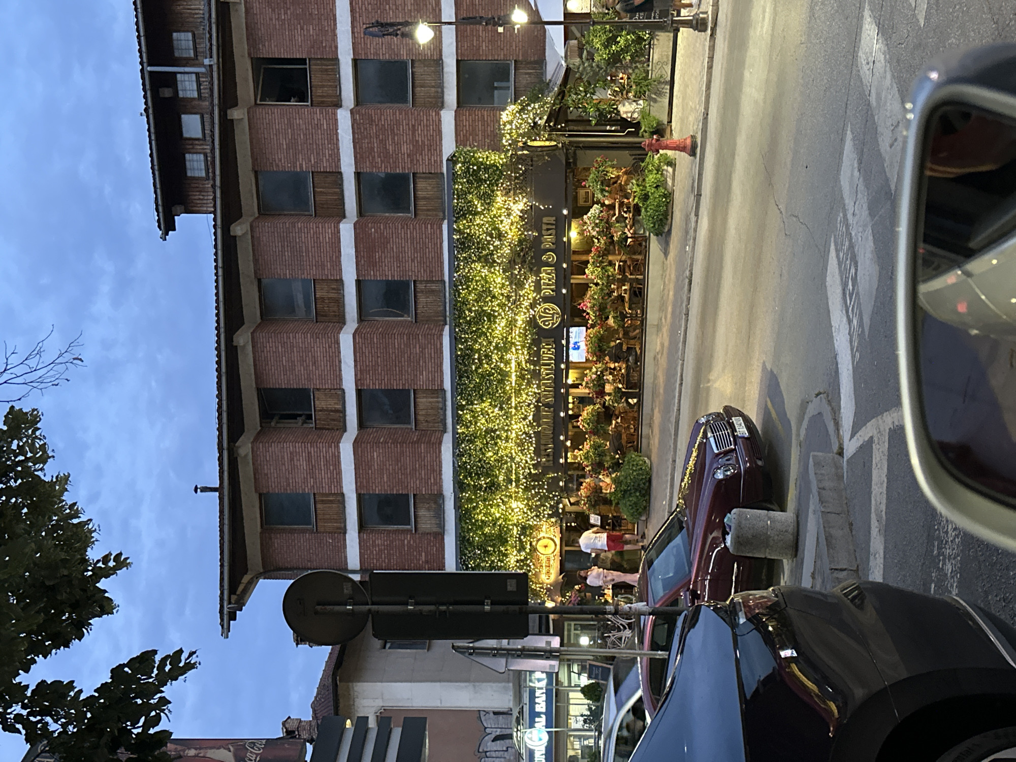 The image depicts a street scene in Veliko Tarnovo, Bulgaria, at dusk. In the foreground, the back of a dark-colored car is visible, as is a car side mirror reflecting a person's face. A maroon Mercedes-Benz sedan is parked on the street to the right of the dark car. In the mid-ground, a building with a red brick facade is adorned with numerous small yellow lights draped over an awning and surrounding plants, creating a festive ambiance. Beneath the lights, a restaurant with the words "PIZZA & PASTA" is visible. Several tables and chairs are set up outside the restaurant, some occupied by diners. A red fire hydrant stands on the sidewalk in front of the restaurant. The street is paved, and a pedestrian crossing is marked. In the background, other buildings and trees are visible under a cloudy sky. The overall impression is of a lively urban evening scene.