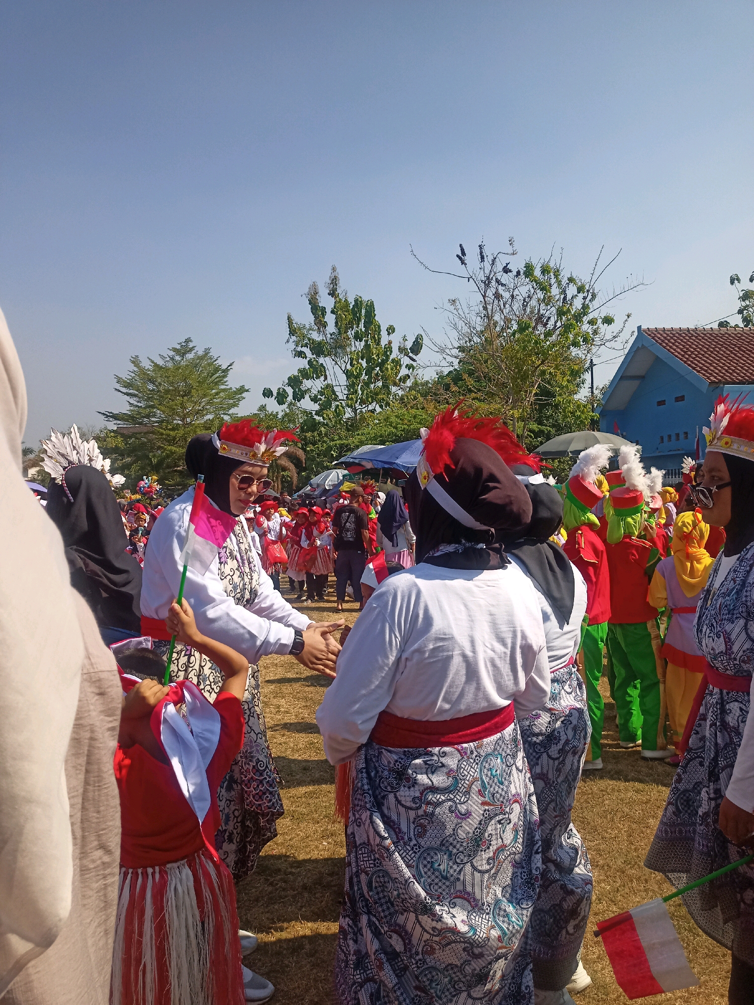 An outdoor event is taking place on a sunny day with a clear blue sky. In the foreground, a woman in a white shirt and a patterned skirt, adorned with a red sash and a feathered headdress, is shaking hands with a child. The child is wearing a red shirt and a white and red fringed skirt, also holding a small Indonesian flag. Behind them, a group of people in colorful costumes, some wearing feathered headdresses and masks, are gathered. In the background, a blue building with a red tiled roof is visible, along with trees and more people in the distance. The ground is covered with dry grass.