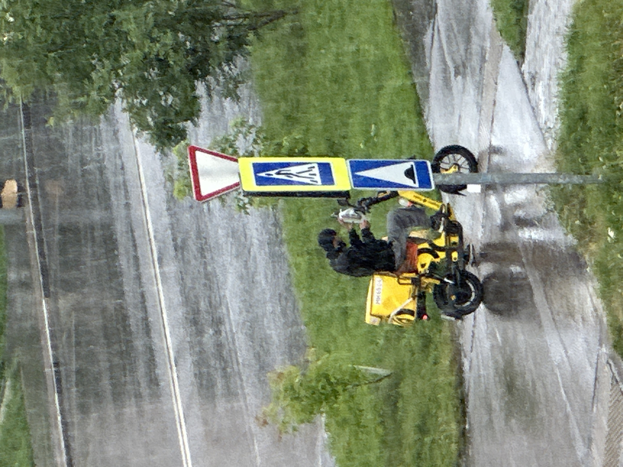 A person wearing a black hooded jacket and gray pants is sitting on a yellow electric scooter in the rain. The scooter has a yellow cargo box on the back with some writing on it. The person is holding a silver object in their right hand, possibly a tool or a phone. They are parked next to a traffic signpost with two signs: a red-bordered yield sign on top and a blue pedestrian crossing sign below it, followed by a blue speed bump warning sign. The scooter is on a wet paved surface, likely a sidewalk or road, which is reflecting the surrounding environment. In the background, there is a tree with green foliage, wet grass, and a road with white lane markings. The overall scene depicts a rainy day in an urban or suburban setting in Podolsk, Russia.