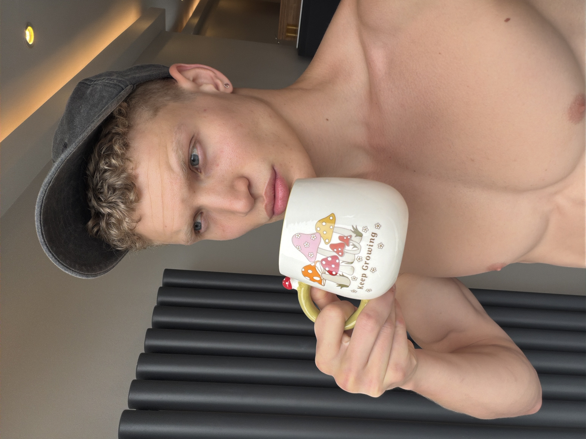 A young Caucasian male with light skin, blonde curly hair, and blue eyes is holding a mug with mushroom illustrations and the words "Keep Growing" printed on it. He is wearing a dark grey baseball cap and no shirt. The background consists of a dark grey radiator and a wall. The image is taken indoors, in what appears to be a residential setting. There is no information in the image to suggest the location is London, United Kingdom.