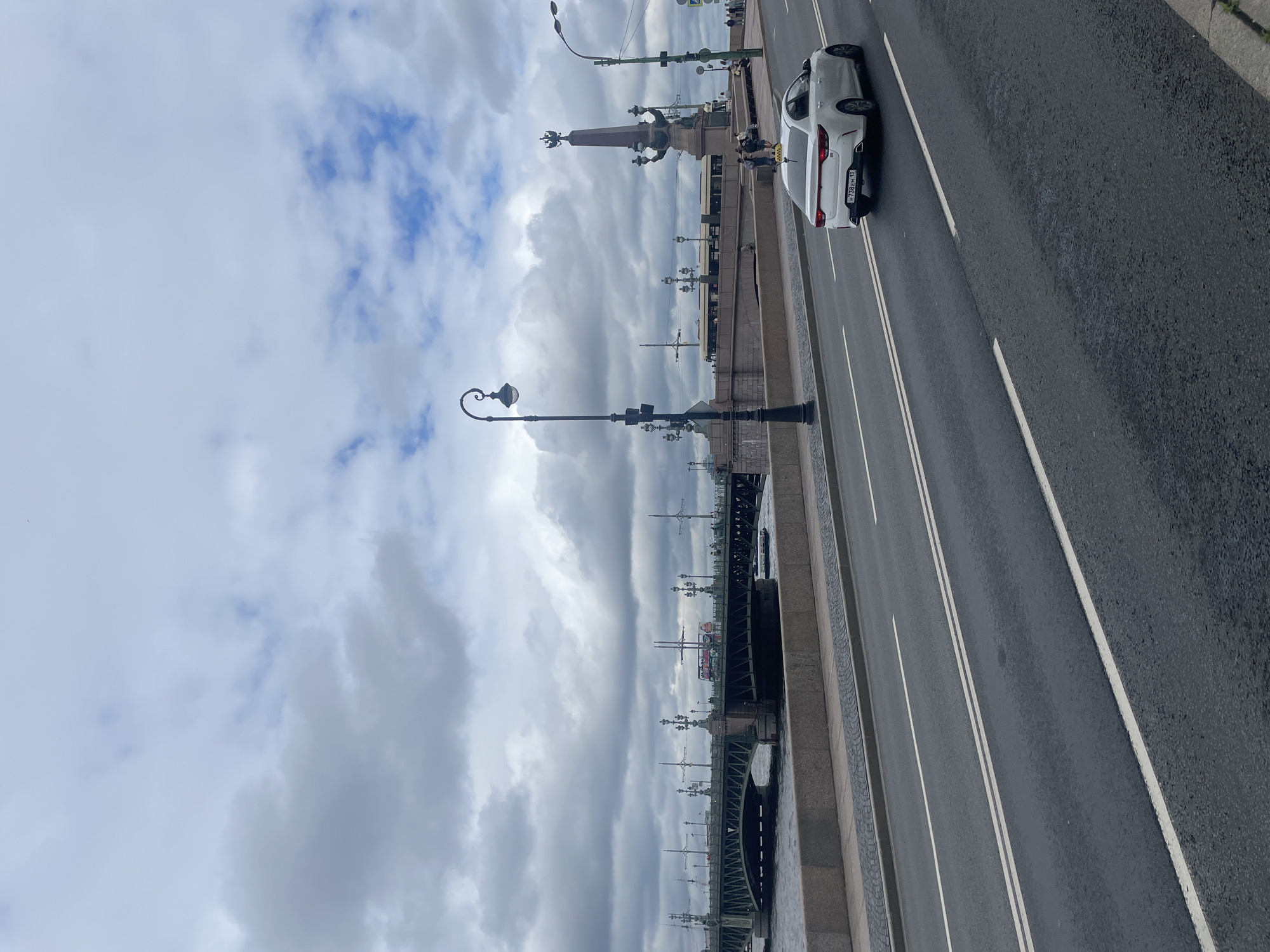The image shows a panoramic view of the Troitsky Bridge in Saint Petersburg, Russia, on a cloudy day. The bridge, a prominent landmark, spans the Neva River and is characterized by its ornate lampposts and the tall Alexandrovskaya column visible in the background. A white Kia sedan is driving on the road leading up to the bridge, with its license plate clearly visible. Several pedestrians are seen walking on the bridge and along the embankment. The Neva River flows beneath the bridge, and a small boat is visible on the water. The sky is filled with dramatic, grey clouds, contributing to the slightly somber atmosphere. The scene captures a typical urban setting in Saint Petersburg, blending historical architecture with modern city life.