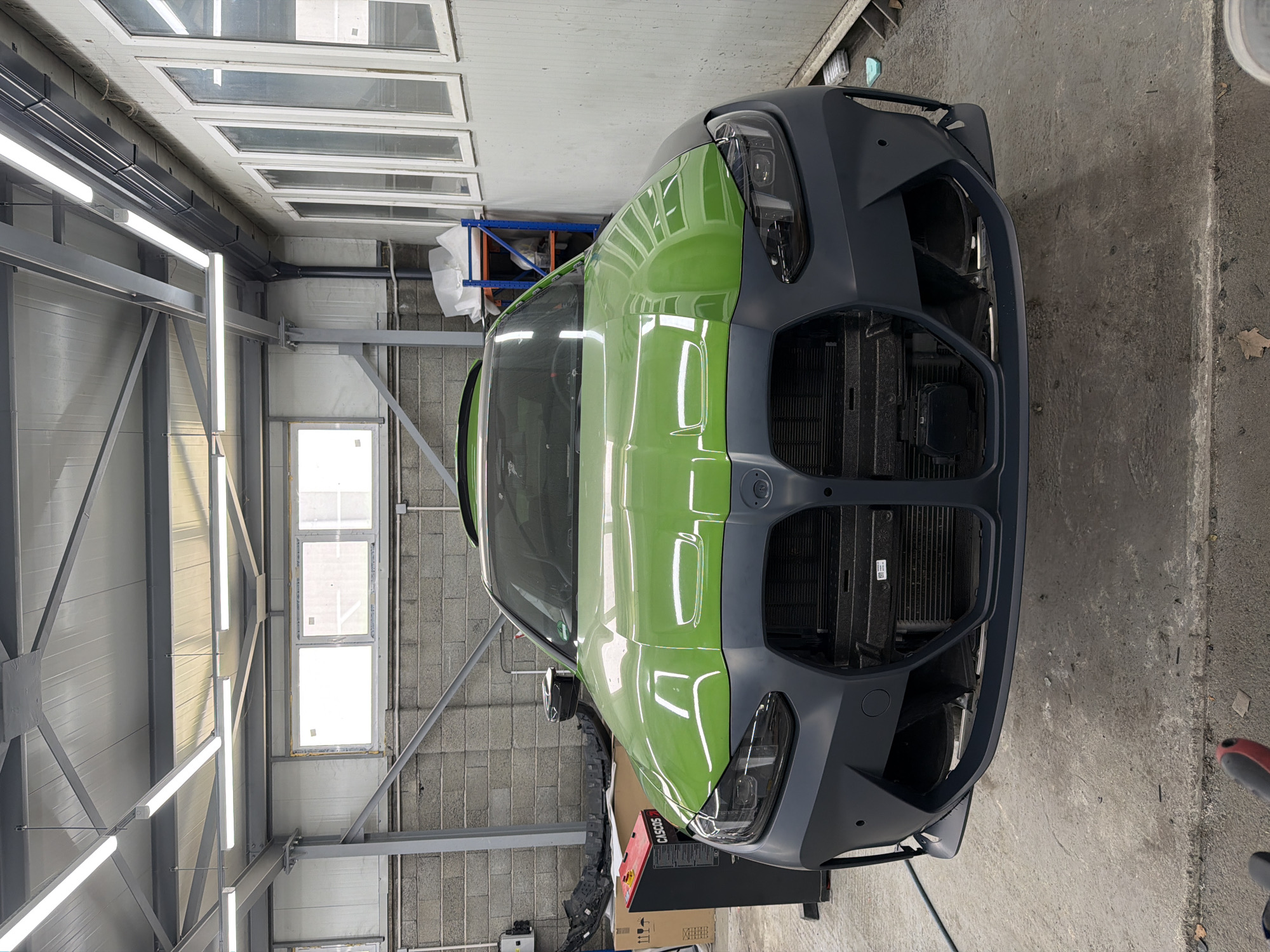 Here's a detailed description of the image:
**Location:** The image appears to be taken inside a large garage or workshop. The specific city and location are unknown.
**Subjects and Objects:**
* The primary subject is the front of a car, likely a BMW based on the emblem. The car's body is a bright green color, while the front bumper and grill are a matte gray/black. The grill is missing, exposing the radiator.
* The environment includes gray metal walls, industrial lighting fixtures, and windows. A metal support structure is visible along the walls. There's a shelving unit with objects covered in plastic sheeting in the background. A red cart and various car parts are situated around the vehicle. The floor is concrete.
**Actions and Interactions:** The car is stationary, presumably undergoing some form of repair or modification. The missing grill indicates that parts have been removed. No people are visible in the frame.
**Environment:** The workshop seems well-lit, due to the industrial lighting. The overall environment is functional and utilitarian, consistent with a space used for automotive work.