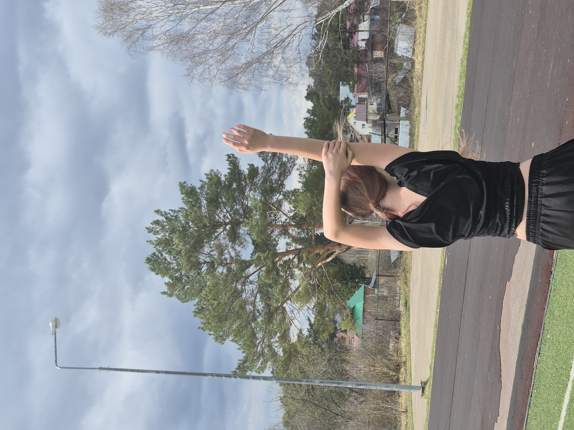 The image captures an outdoor scene at what appears to be a sports facility in Chaikovsky, Russia, under an overcast sky. The central subject is a person, seen from the waist up and from behind, performing an overhead arm stretch. They have light brown hair tied back and are wearing a black short-sleeved athletic top with a cinched waist, and black shorts or pants. Their arms are extended upwards, with one hand gripping the opposite forearm or wrist.
The setting includes a dark asphalt track surface and an adjacent strip of green artificial turf on the right side of the frame. On the left, a tall metal street light pole with a luminaire extends into the sky. The background is dominated by a large, lush green coniferous tree, possibly a pine. Bare deciduous trees are also visible, indicating either early spring or late autumn. Beyond the trees, a cluster of residential houses with various colored roofs can be seen. The sky is filled with light grey, unbroken clouds, suggesting a daytime scene without direct sunlight. There is no visible text in the image.
