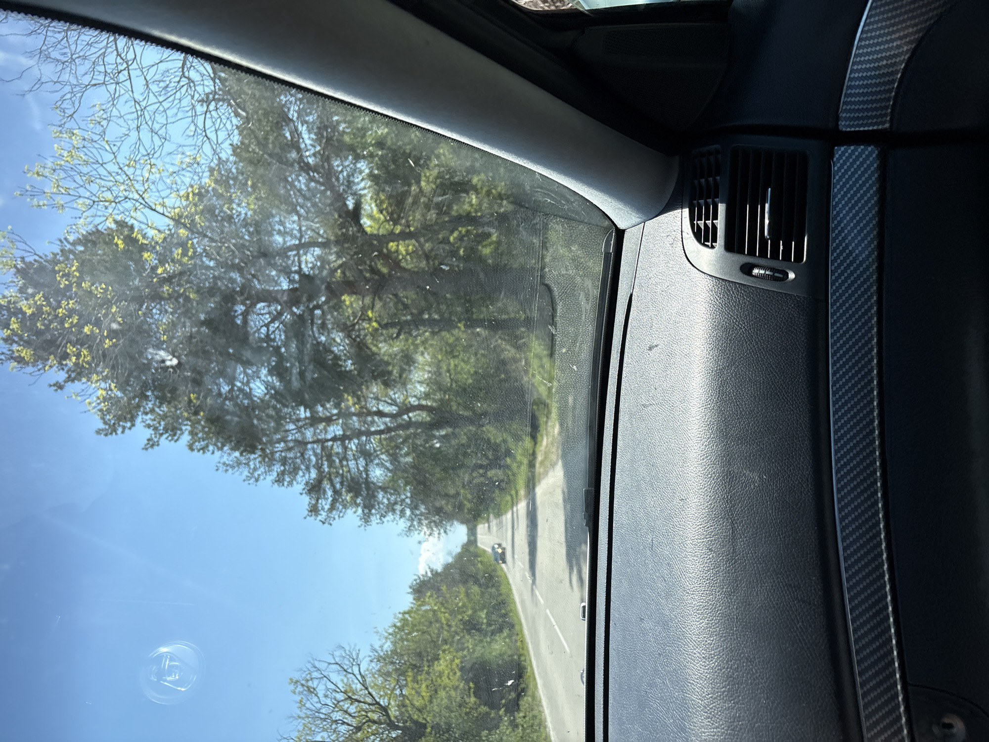 The image depicts a view from inside a vehicle traveling on a road. The exterior scenery, visible through the passenger-side window, shows a winding road lined with trees under a clear, blue sky. The vehicle's dashboard is partially visible on the right side of the frame, featuring an air vent and textured material. The precise location within the city of Levski, Bulgaria, cannot be determined from the visual information provided. The image does not show any people.