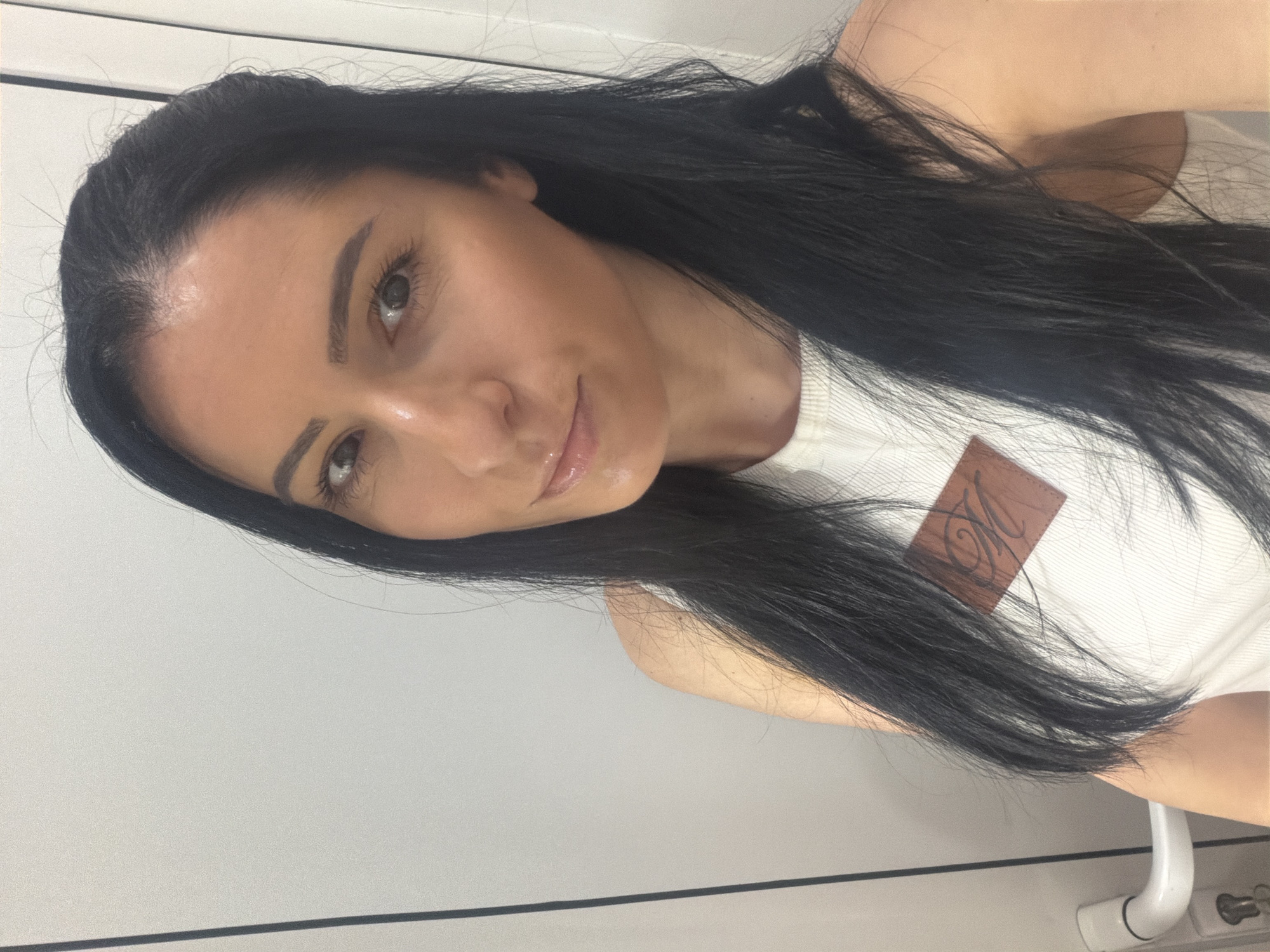One female subject is depicted in a close-up, frontal portrait, situated in the city of Pleven, Bulgaria. She has long, straight, dark hair, a fair complexion, and her dark eyes engage directly with the camera. The subject wears a ribbed, light-colored sleeveless garment. A brown, rectangular patch displaying a stylized 'M' or similar emblem is affixed to the upper left chest area of her attire. The background consists of a light-colored vertical surface, identified as a door, evidenced by a partially visible white lever door handle and a keyhole on the lower left side of the frame.