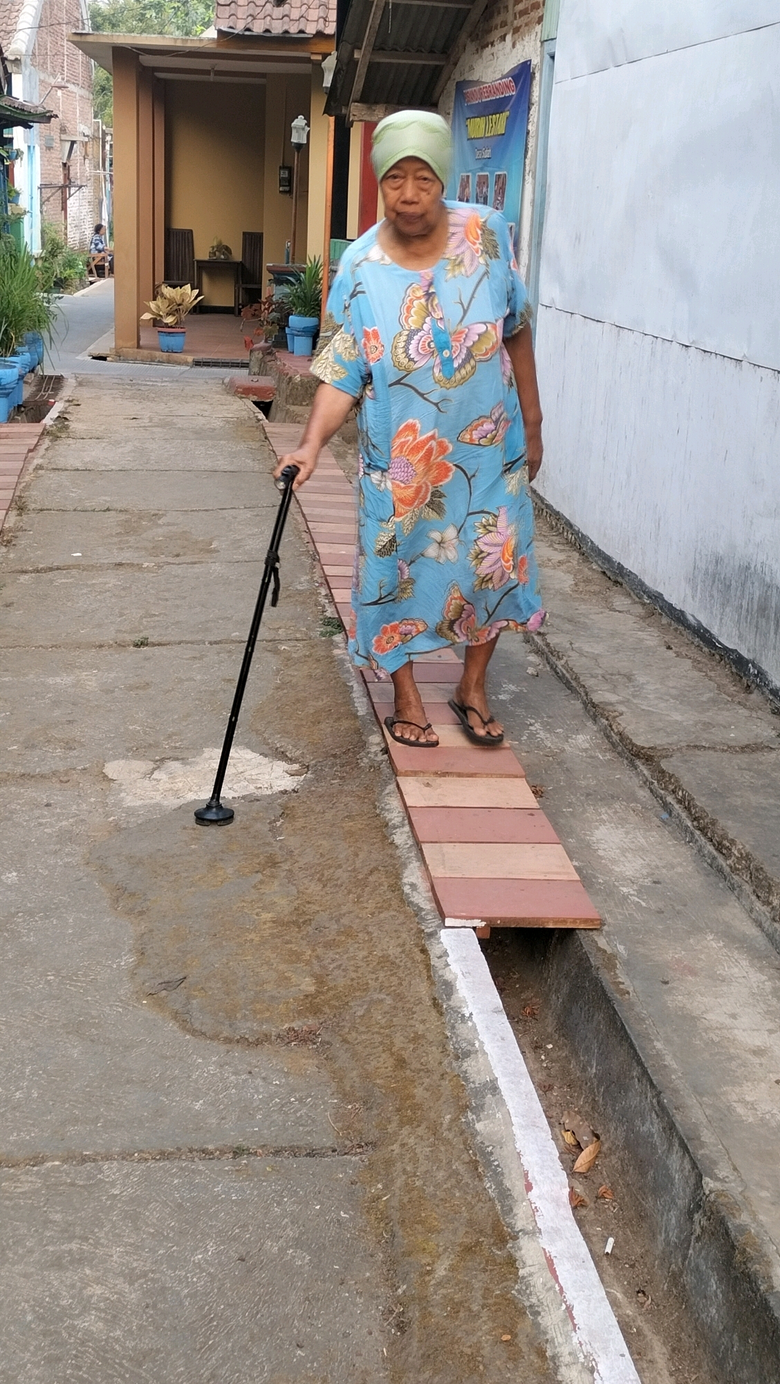 An elderly Indonesian woman, wearing a light blue dress with a floral pattern and a green headscarf, stands on a narrow wooden bridge. She is holding a black cane with a wide base. The bridge spans a drainage ditch and is constructed from several planks laid across it. The woman appears to be walking across the bridge. The environment is an outdoor urban setting, likely in Subah, Indonesia, with buildings and a wall visible in the background. The ground around the ditch is concrete, with some dirt and debris.