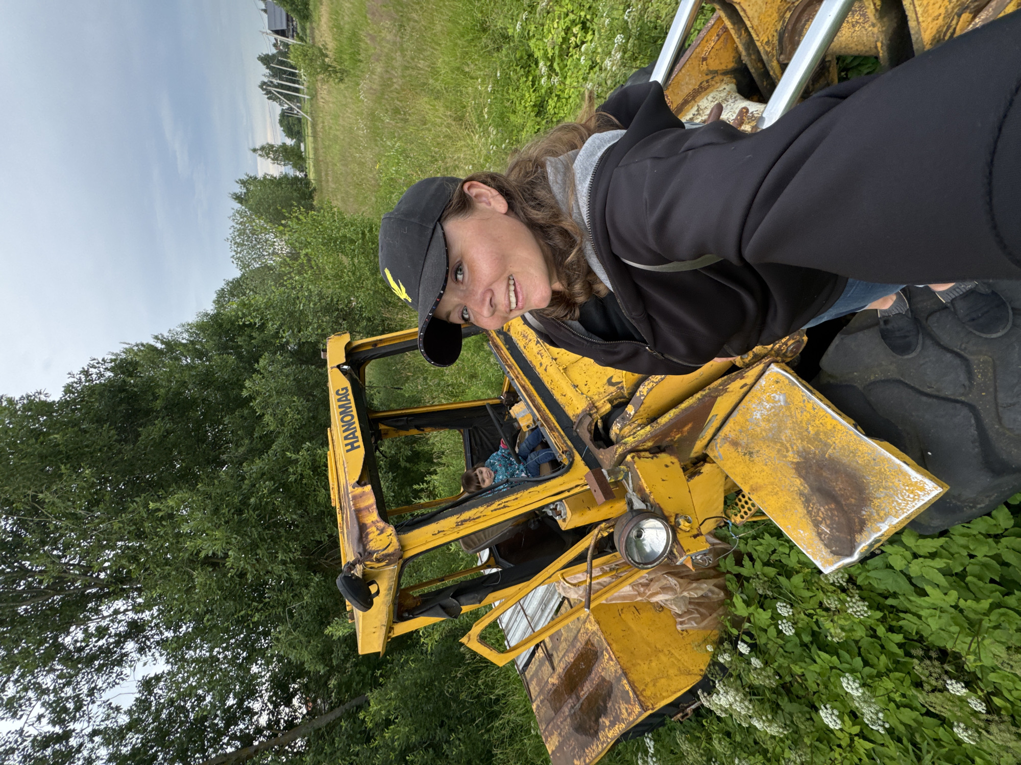 This is a medium shot photograph taken in Innolovo, Russia, on a cloudy day. A woman in a black baseball cap and black zip-up hoodie is standing next to an old, rusty yellow tractor, holding the camera for a selfie. She is smiling at the camera. Inside the tractor's cab, a young child is sitting in the driver's seat. The tractor is situated in a grassy area with abundant green foliage and small white flowers. Trees are visible in the background, and a village with houses and utility poles can be seen in the distance. The overall scene depicts an outdoor, rural setting with a focus on the woman and the child interacting with the abandoned tractor.