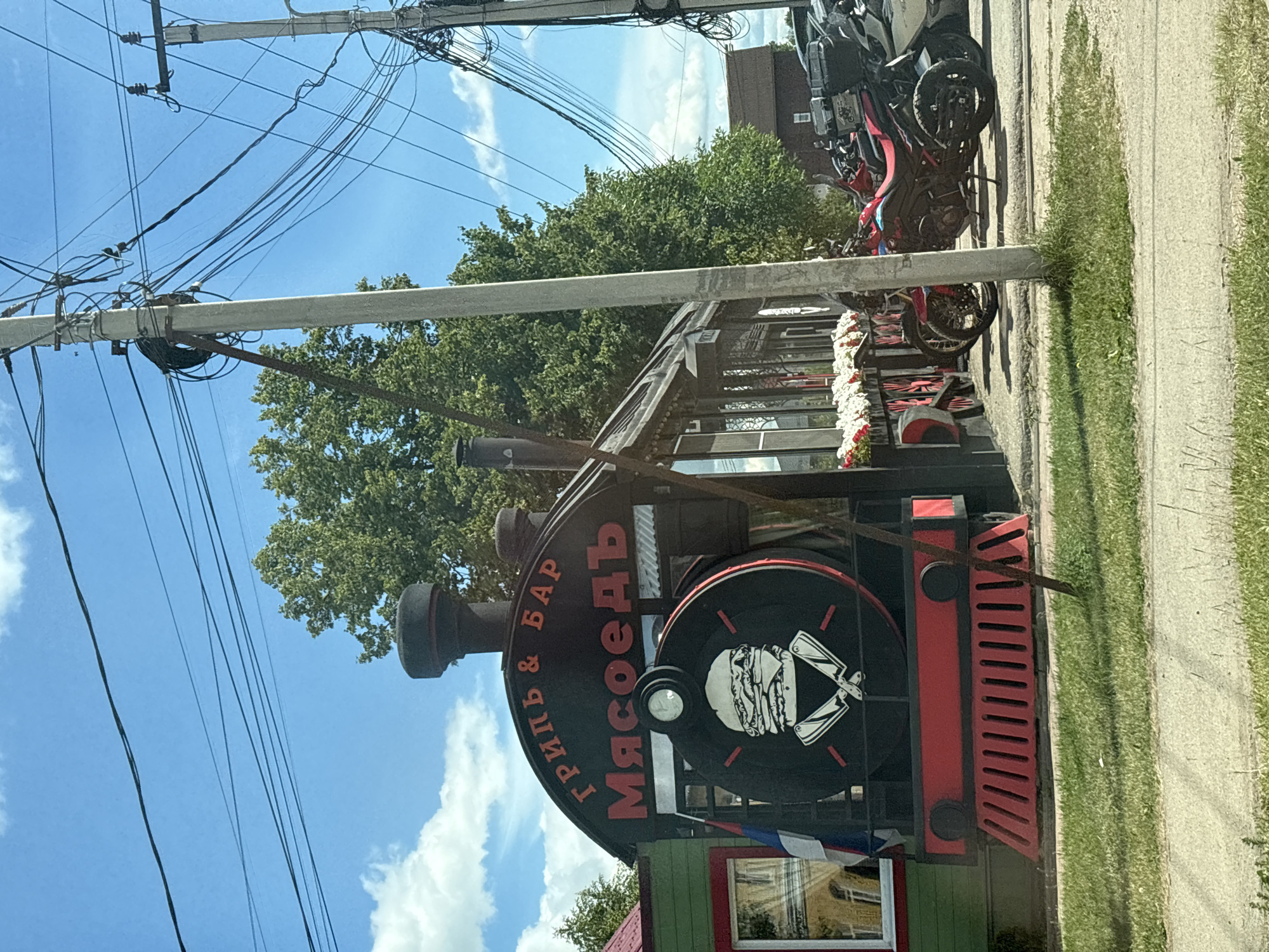 The image depicts the exterior of a restaurant designed to resemble a train. The restaurant, identified by the Cyrillic text "ГРИЛЬ & БАР Мясоедъ" (Grill & Bar Myasoyed) on its facade, is located in the city of Pereslavl-Zalessky, Russia. The front of the building is stylized as the engine of a steam locomotive, complete with a circular emblem featuring a stylized burger and two crossed knives. Red decorative elements, resembling the train's wheels and grill, are visible at the base. The restaurant is flanked by a green building on the left, and a row of motorcycles and a car are parked in front of it on the right. The scene is set against a backdrop of trees and a blue sky with scattered clouds. Electrical poles and overhead wires are present in the foreground and midground. The overall impression is of a unique, themed dining establishment in a suburban or roadside setting.