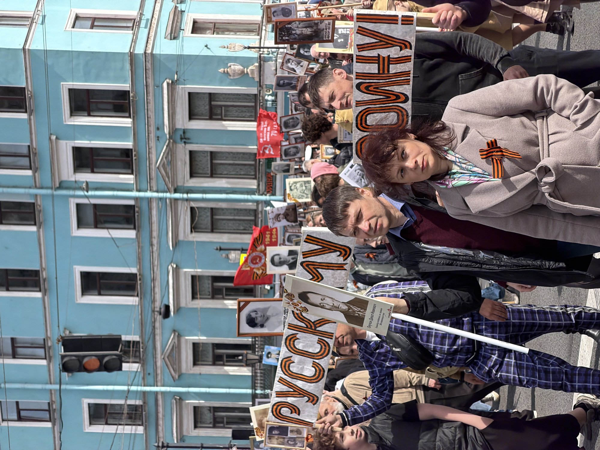 A crowd of people marches on a street in what appears to be Saint Petersburg, Russia. They are holding portraits and banners with text in Russian, and some individuals wear ribbons with orange and black stripes. The architecture of the buildings in the background features light blue facades and detailed window frames. A traffic light is visible near the buildings. The people seem to be engaged in a procession or a public gathering.