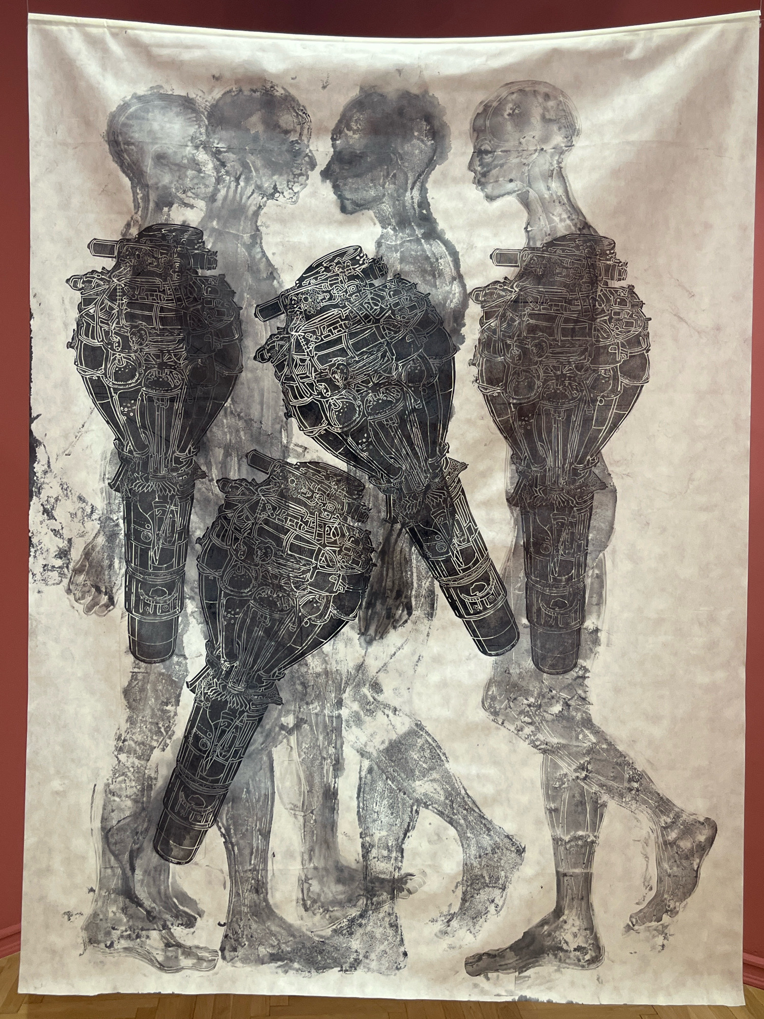 This is a black and white, full-shot, eye-level photograph of a large, abstract artwork displayed on a pale beige wall with a reddish-brown border. The artwork, which appears to be a print or painting on fabric, depicts several overlapping, semi-transparent figures. The figures are rendered in shades of grey and black, with some areas showing more detail than others.
In the foreground and dominating the composition are two stylized, mechanical-looking appendages that resemble cannons or complex engine parts. These are intricately detailed with lines and circular elements, and are positioned in the lower half of the artwork, one slightly in front of the other.
Behind these mechanical elements, the forms of human figures are visible. The heads and upper torsos of at least four figures can be discerned, layered and faded, suggesting movement or a ghostly presence. Their features are indistinct, and their bodies merge into the background. The lower bodies and legs of some figures are more clearly rendered, showing the form of feet and legs in motion. The overall impression is one of a blend of the organic and the mechanical, with a sense of fragmentation and superposition.
The artwork is illuminated by ambient lighting, casting soft shadows. The wall it is mounted on is a plain, matte surface. The bottom edge of the artwork extends to the floor, where a polished wooden surface is visible. The reddish-brown border suggests the artwork is either framed by a wall of that color or is a large tapestry with a colored border.
The specific location within Podgorica, Montenegro is not discernible from the image itself.