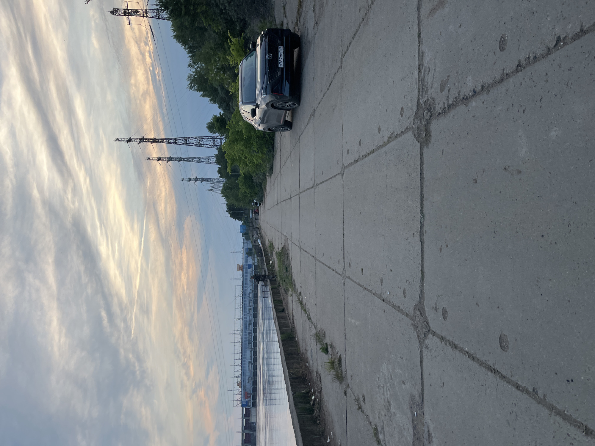The image depicts an expansive outdoor scene, likely an embankment alongside a large body of water, under a late afternoon or early evening sky. A straight concrete path, constructed from worn slabs with minor cracks, runs along the right side of the frame. A dark-colored SUV, possibly a Kia, is parked facing away from the viewer on this path.
To the left of the path, a calm body of water extends into the distance, bordered by a substantial, possibly industrial or dam-like structure on the far bank. Multiple tall metal power line towers (pylons) punctuate the landscape, appearing both along the water's edge and further inland, suggesting a significant electrical infrastructure. Green trees line the area behind the concrete path and along the opposite bank of the water.
The sky exhibits soft, wispy clouds with patches of blue, illuminated by warm, golden light characteristic of low sun. The overall scene is static, with no human activity visible. The license plate on the parked SUV reads "О787МО 152". The presence of the large water body and extensive power grid aligns with the location context of Chaikovsky, Russia, home to the Votkinsk Hydroelectric Power Station.